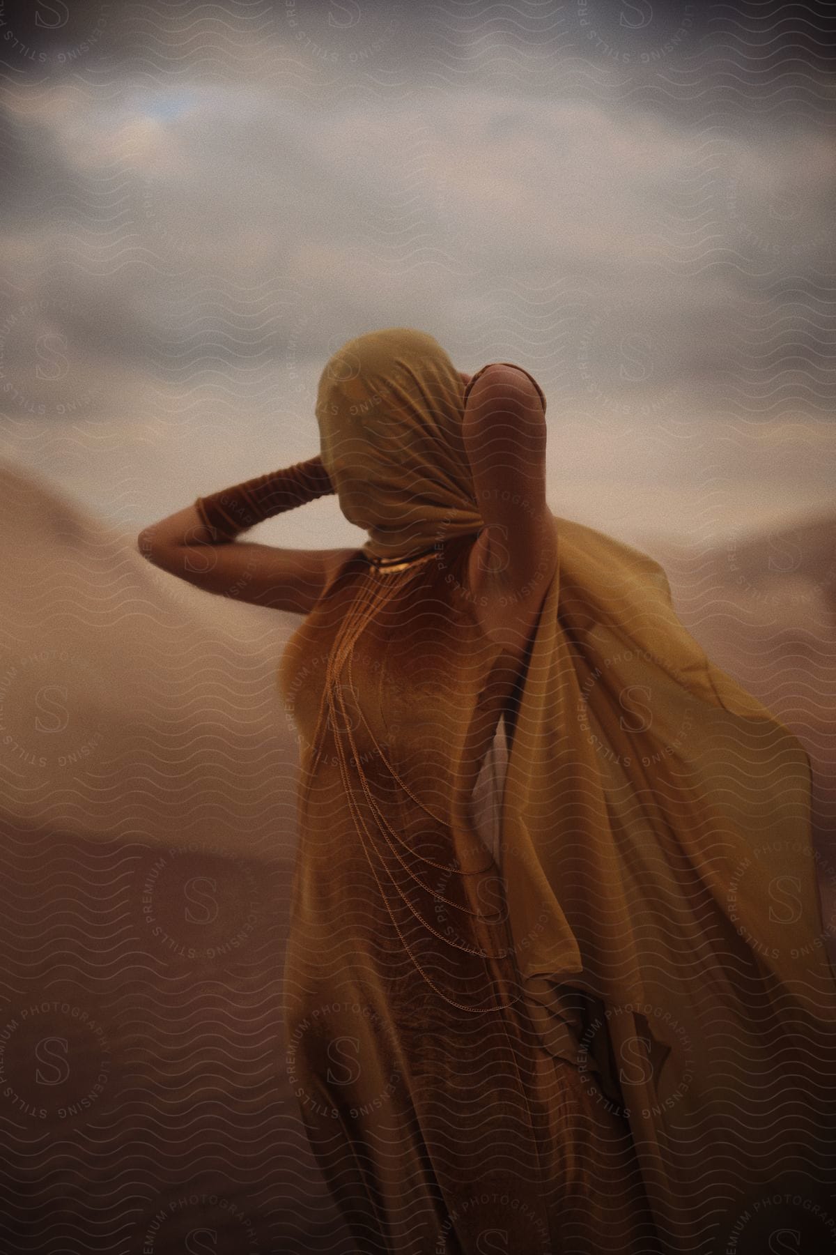 Amidst a blustery, overcast day, a woman in a brown dress shields her face from the elements, fastening a brown scarf around her head as she stands alone in the vast desert.