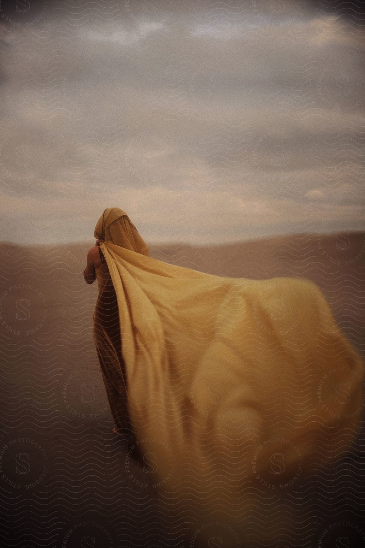 Woman Wearing A Gown With A Long Hooded Cloak Walks Across Sand On A Windy Overcast Day