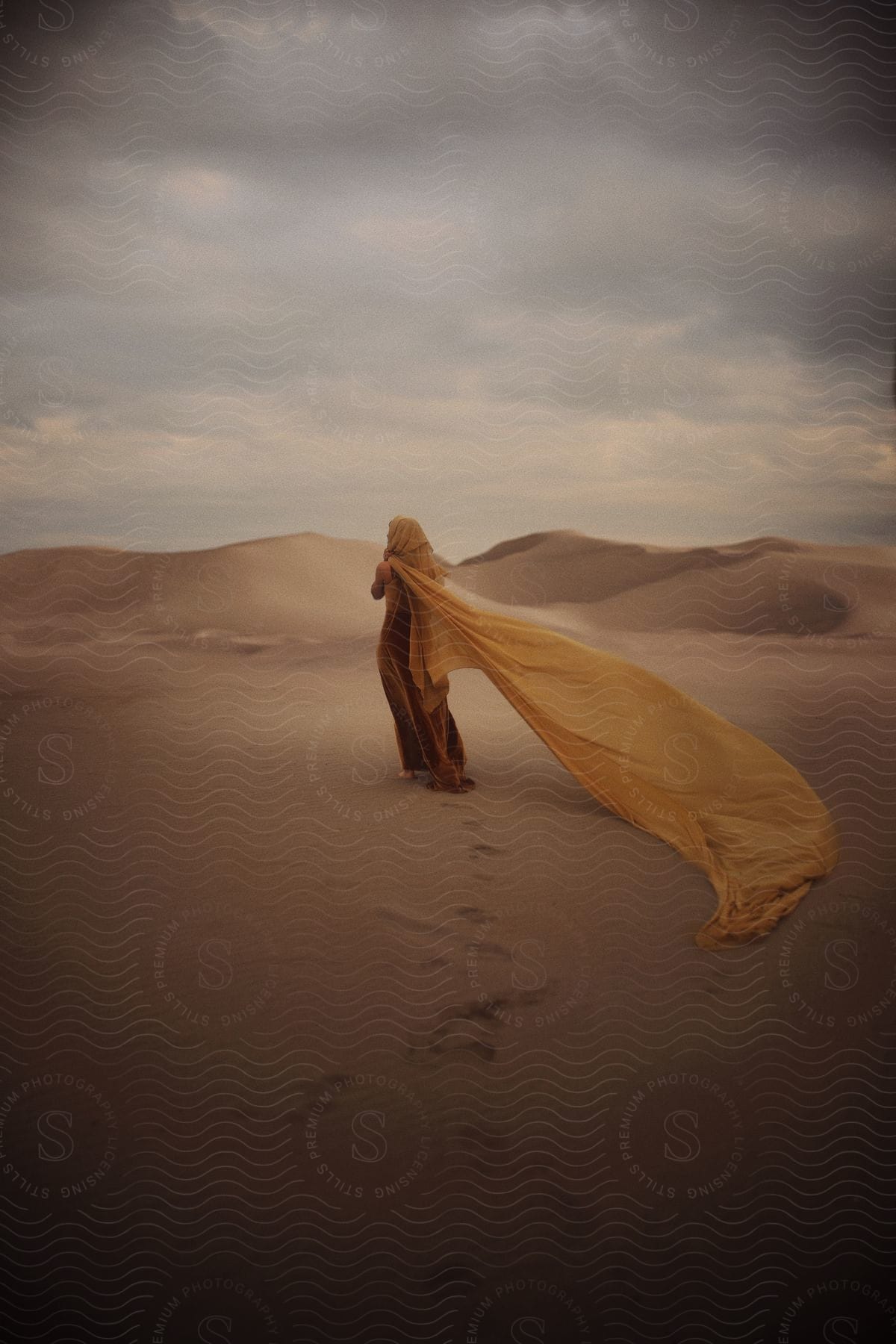 A Person Is In The Desert, Wrapped In A Long Flowing Yellow Fabric That Flutters In The Wind