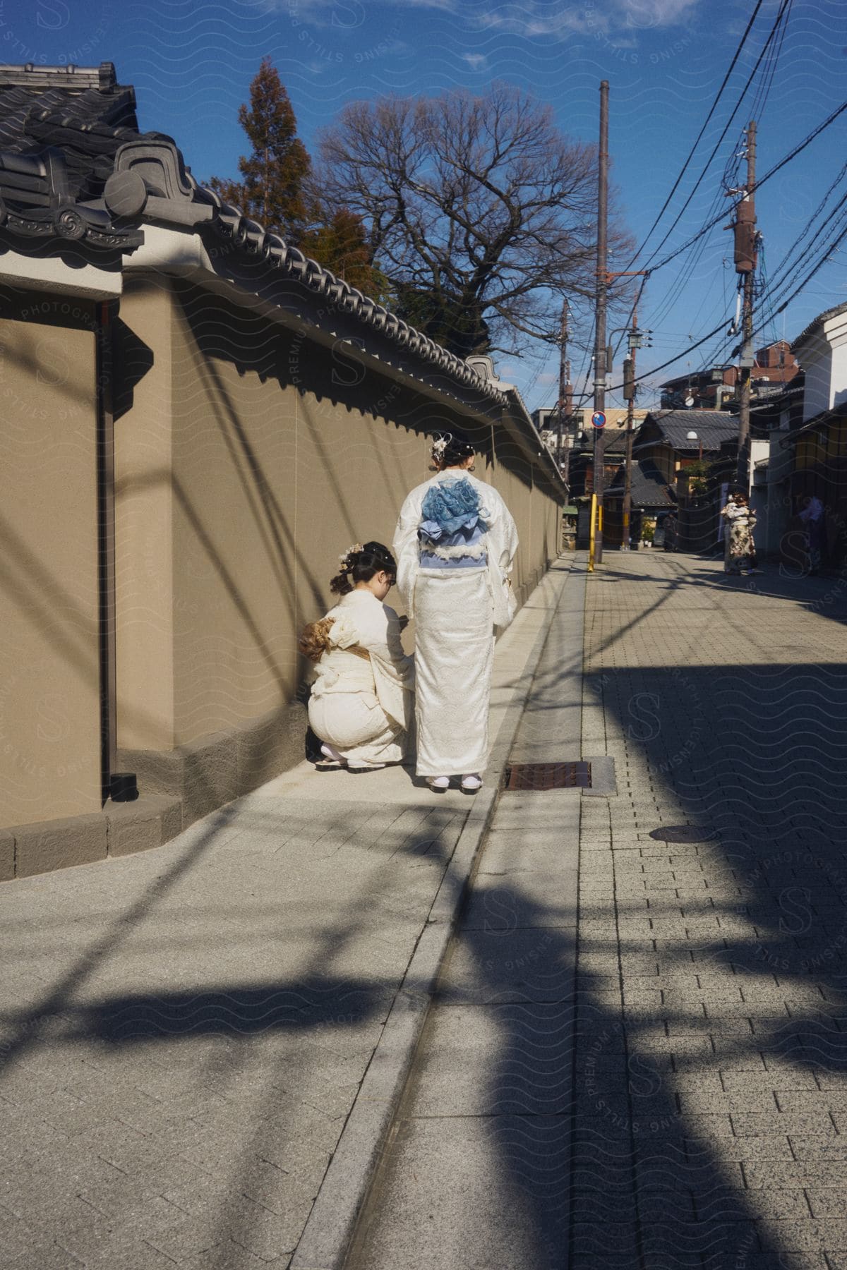 Two people are on the sidewalk, one kneeling and the other standing, both wearing traditional dress. The sky is clear and blue, indicating a sunny day.