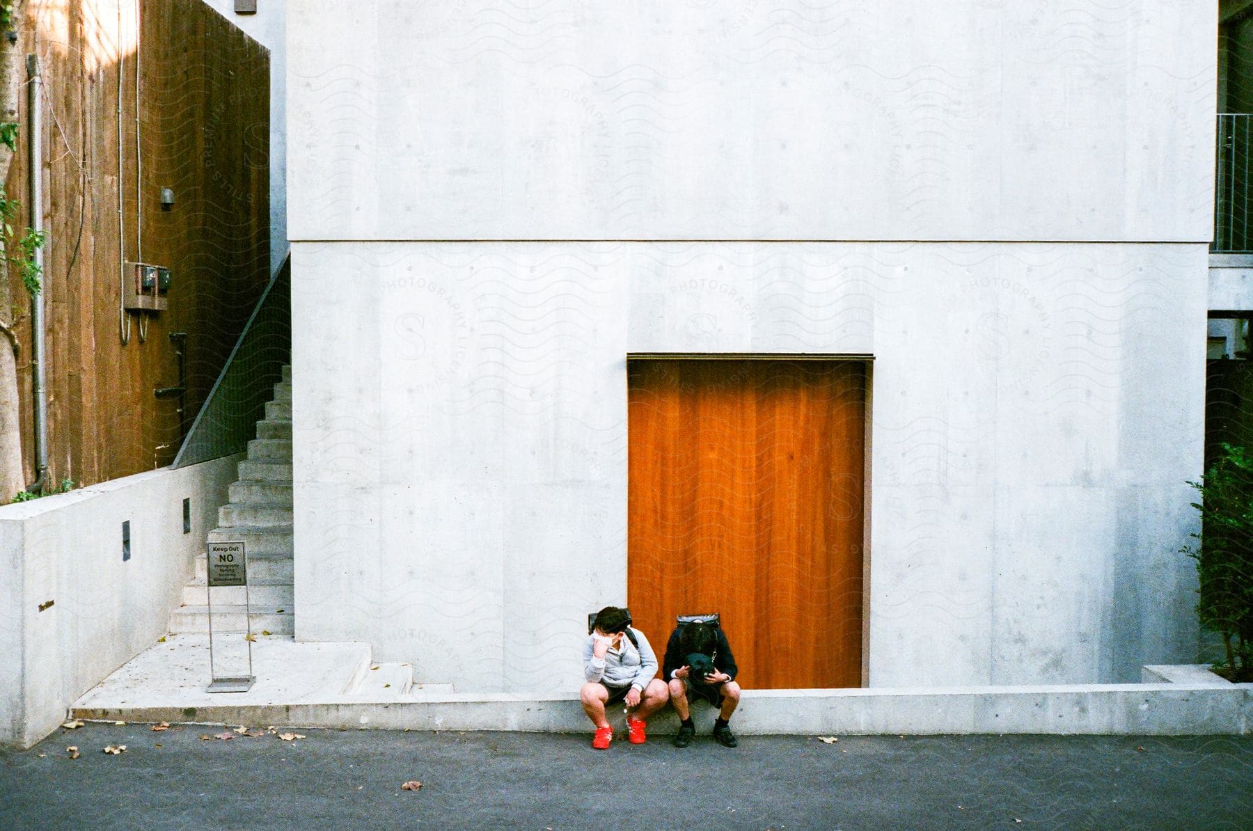 Two schoolboys, backpacks slung over their shoulders, perch atop a cement barrier beside a vibrant orange door, with a "No Entry" sign prominently displayed in front of a staircase to the left.