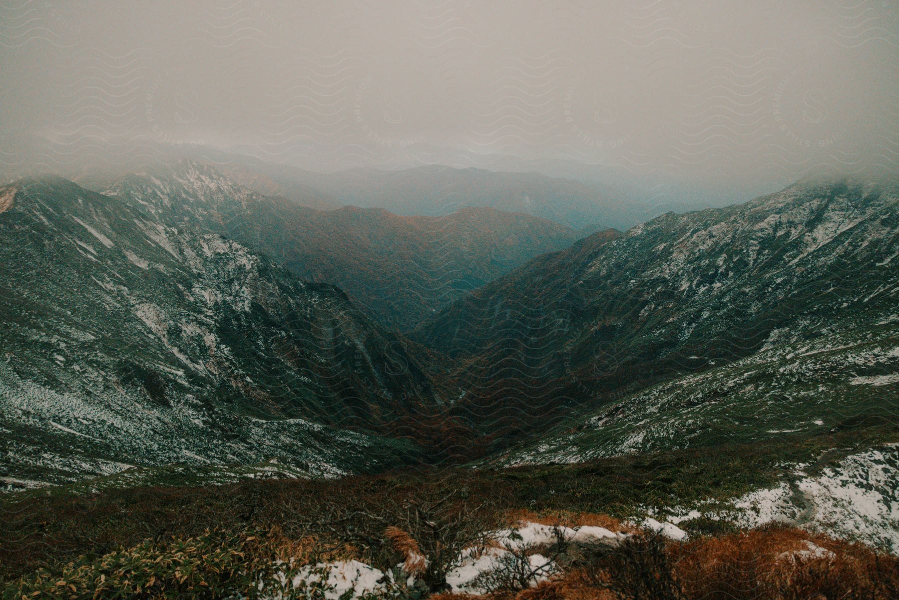 During the fall season, mist covers the sky around a mountain with a sloping descent, and patches of snow are visible at its summit.