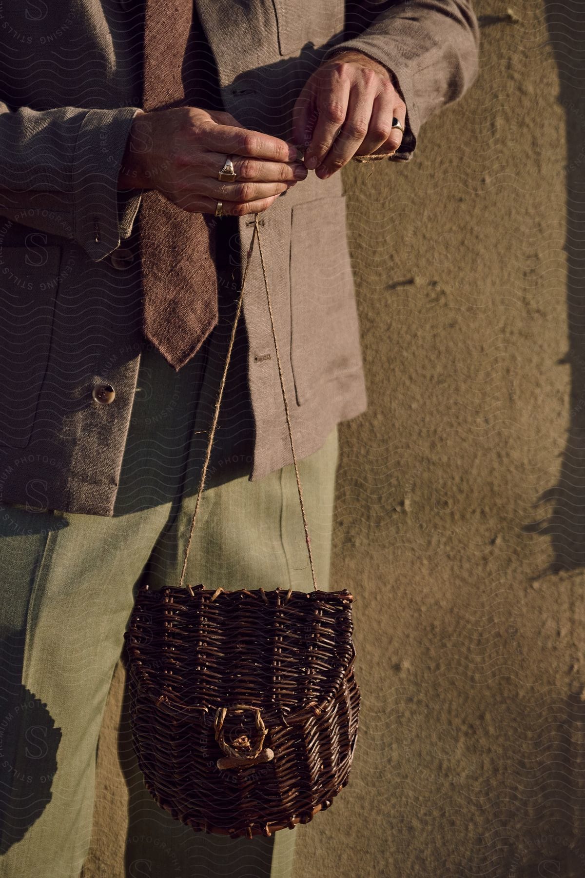 Close-up of a person's midsection with hands fastening a brown wicker bag; wearing a grey suit jacket, brown vest, and green trousers against a textured wall in daylight.