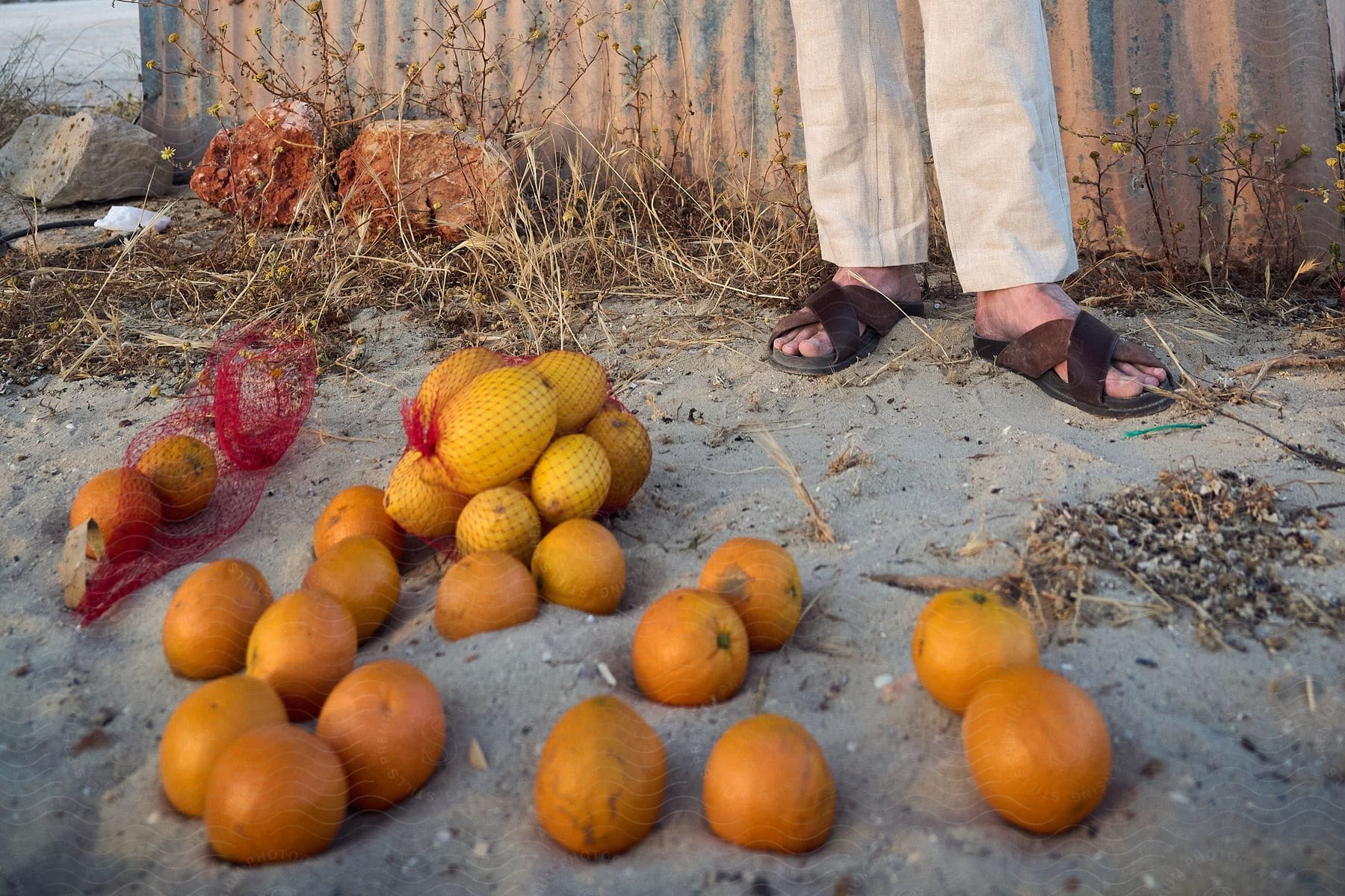 Bag with some oranges thrown on the sand floor and in the background a person's feet.