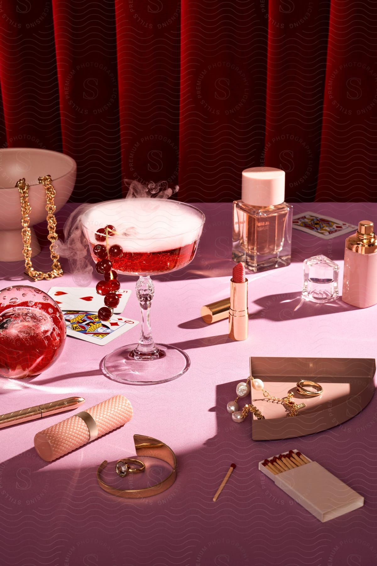 Pink table with red cherry cocktail and jewelry and makeup around it.