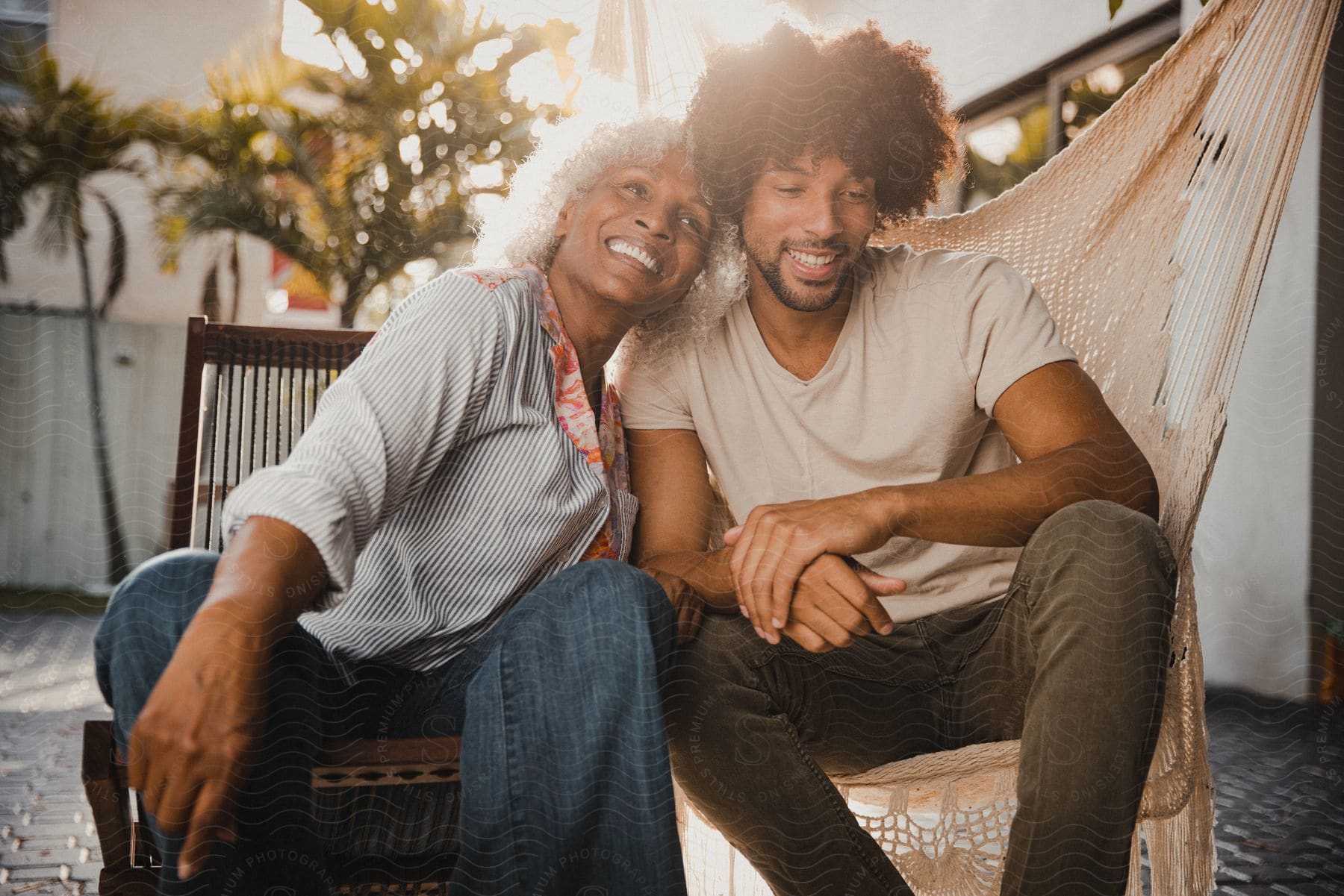 Mother sitting in a chair next to her son who is sitting in a hammock while she rests her face on his shoulder and they both smile happily.