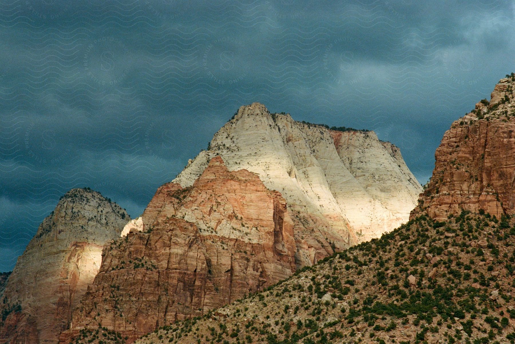 Green shrubs grow on a mountainside beneath a dark, cloudy sky in a desert mountain range.