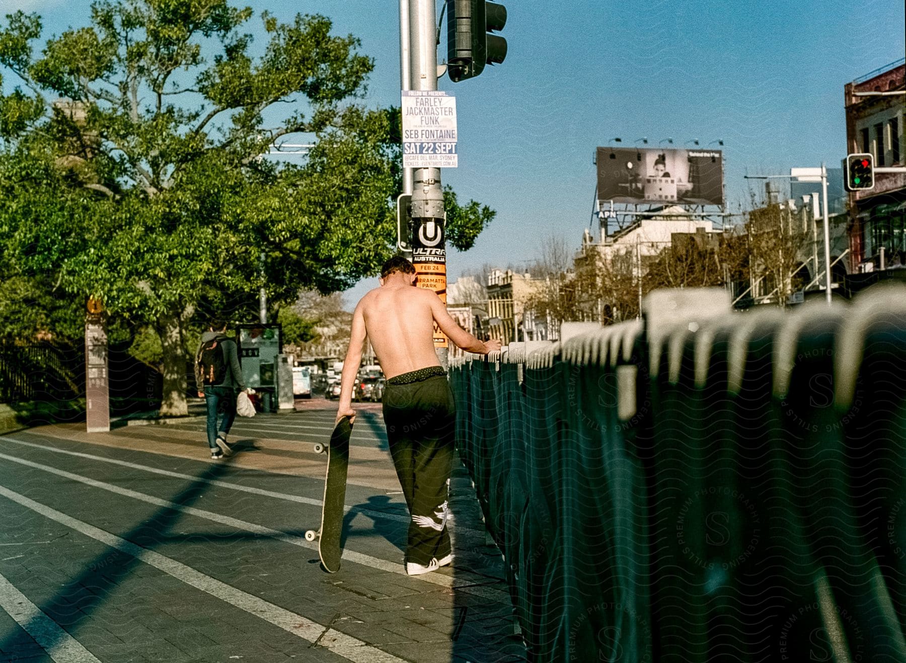 A shirtless man with a skateboard walks on a crosswalk by a traffic light and a fence in a suburban area.