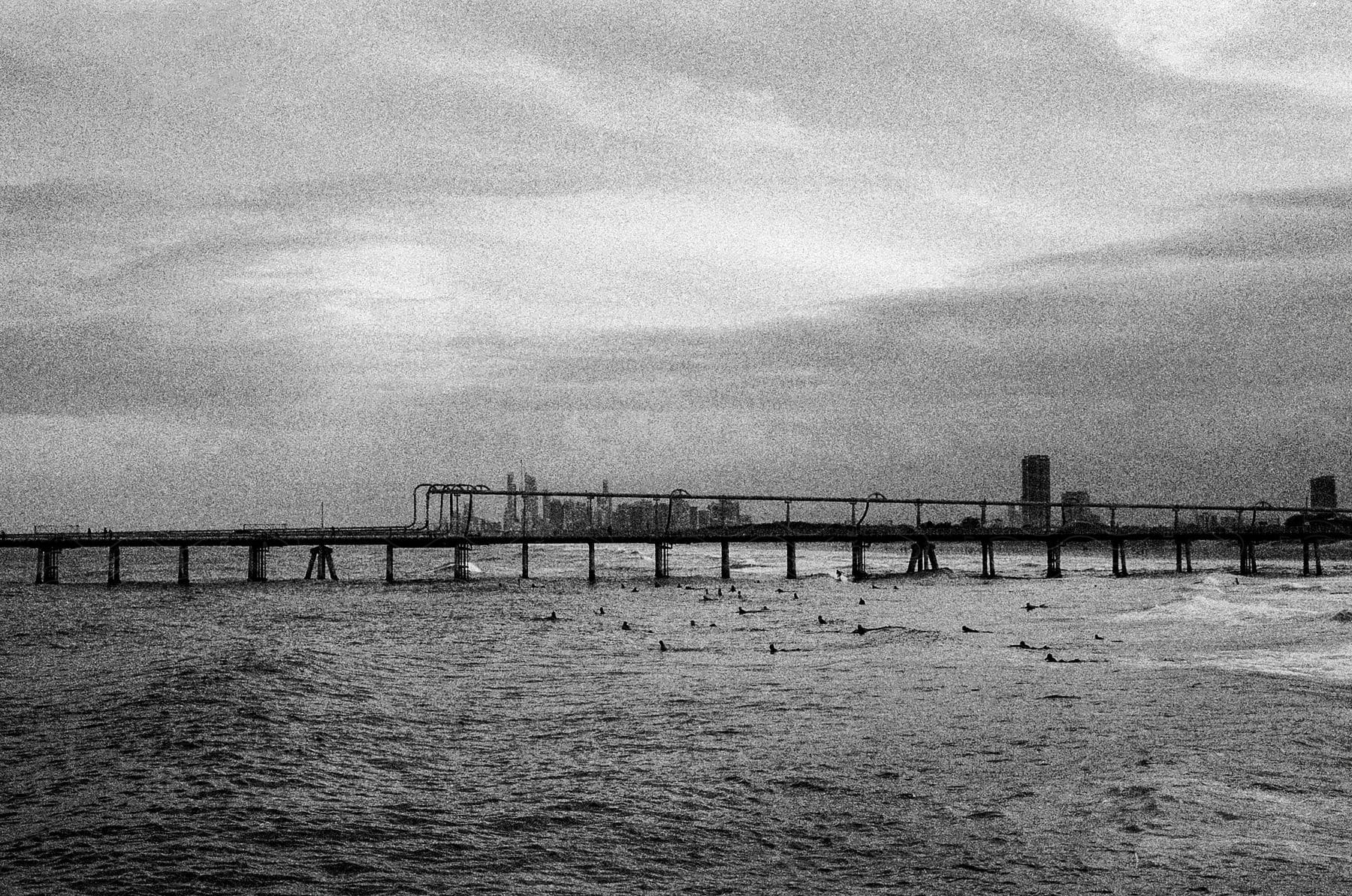 People swim in water near silhouetted pier a distance from skyline on a cloudy day.