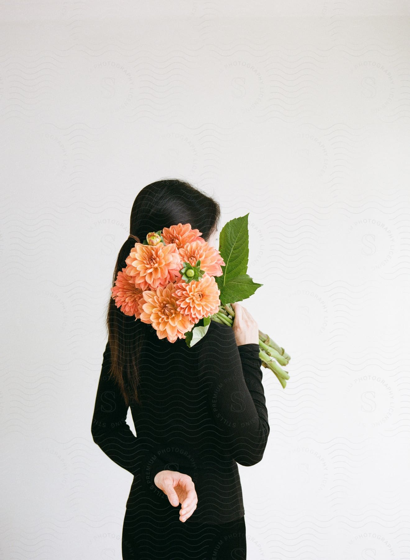 A woman dressed in black, with one hand behind her back and a flower bouquet over her shoulder.