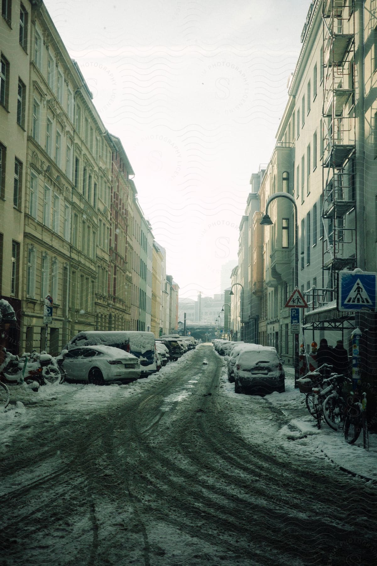 Snow-covered street with parked cars and bicycles in an urban setting, flanked by European-style buildings