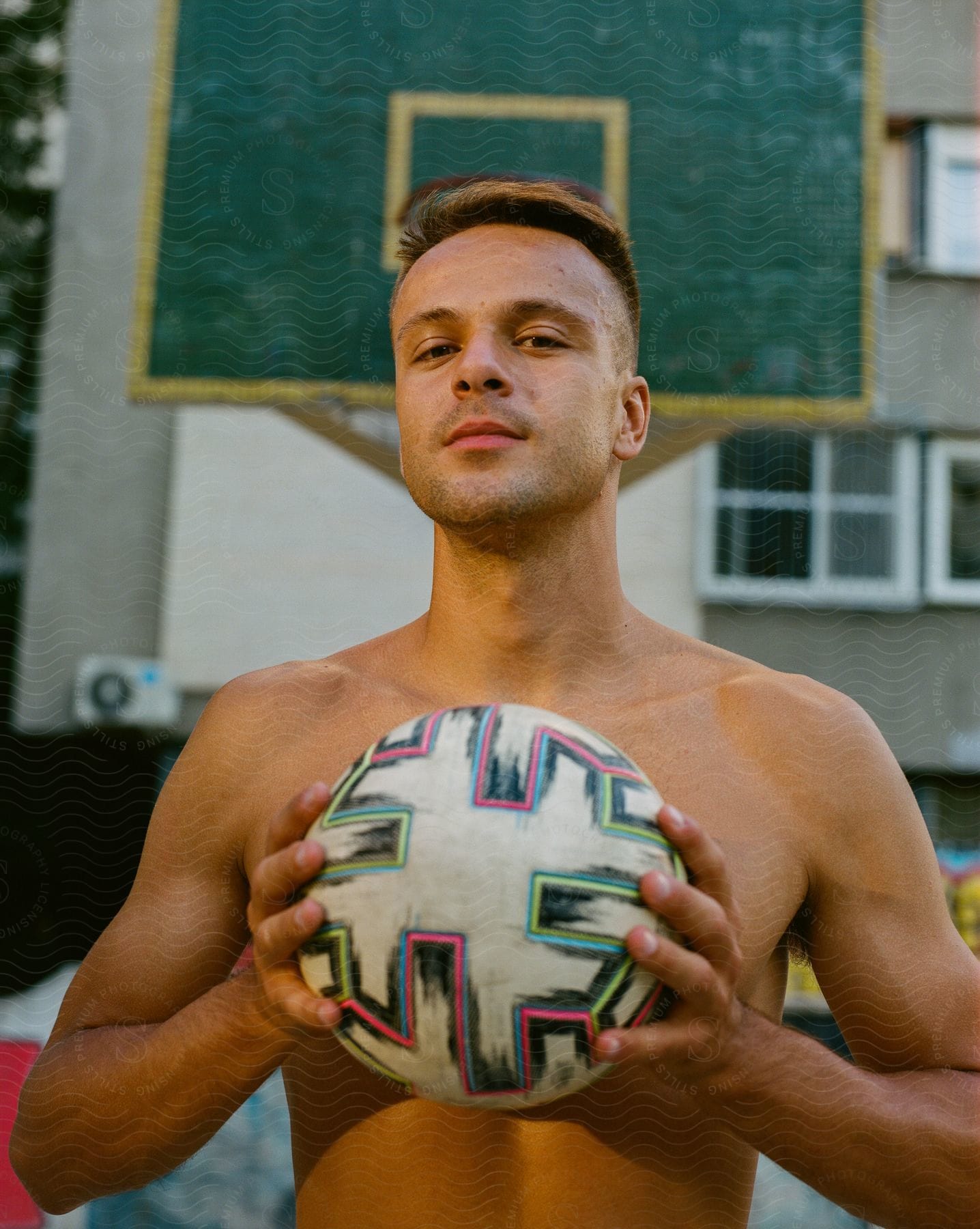 Shirtless man posing with a soccer ball outdoors.