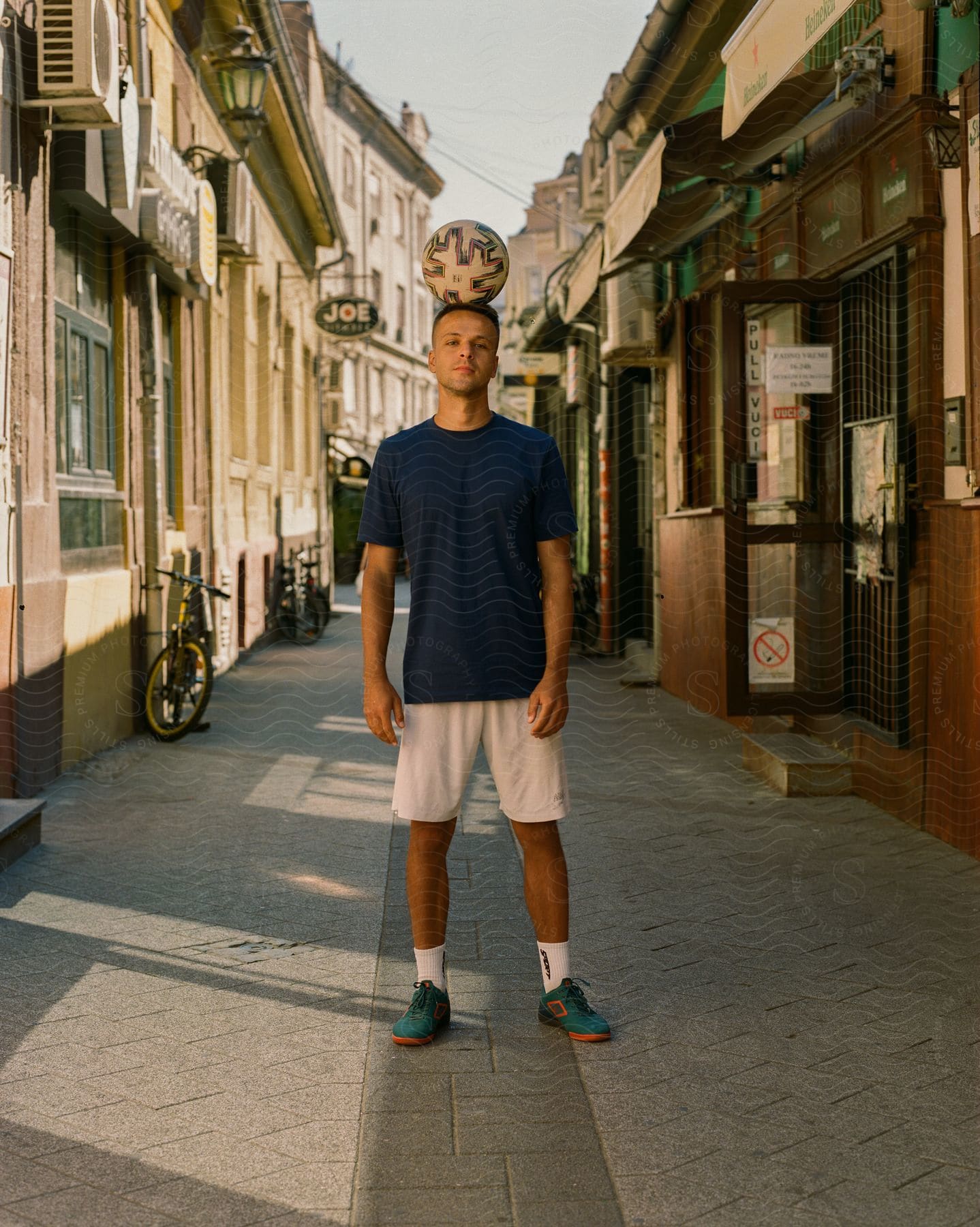 A man wearing a navy T-shirt, white shorts, socks, and green shoes stands in the narrow downtown area.