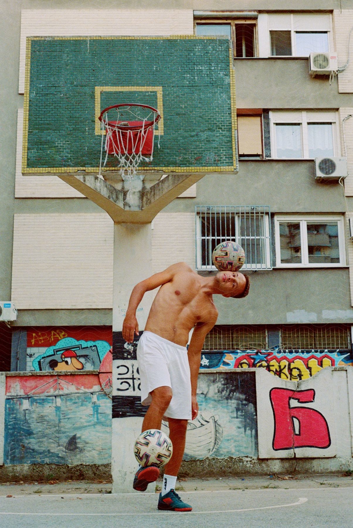 A shirtless person balances a soccer ball on their head in front of a graffiti-covered wall and a weathered basketball hoop.