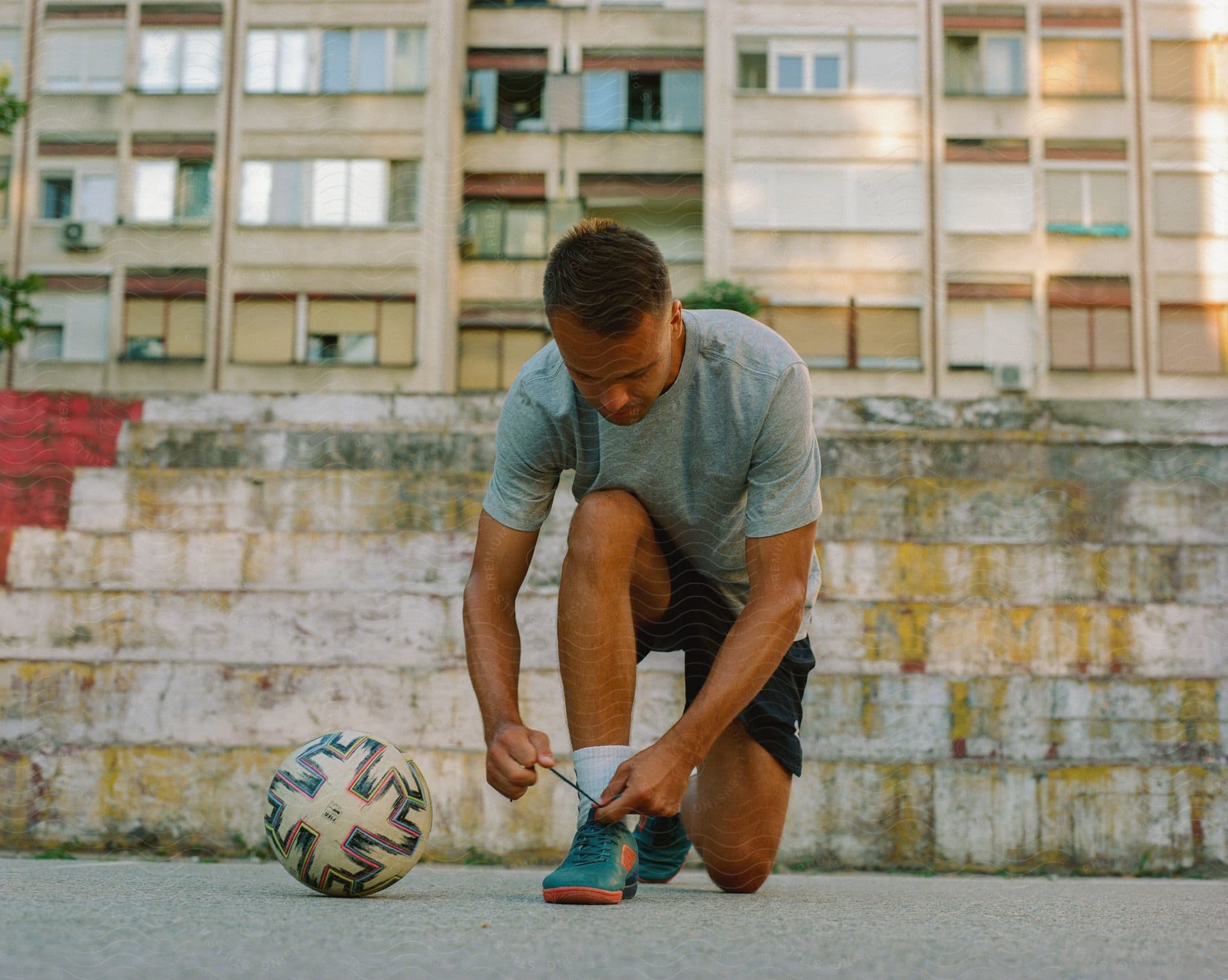 A person is tying their shoe next to a soccer ball on a street with a multi-story building in the background.