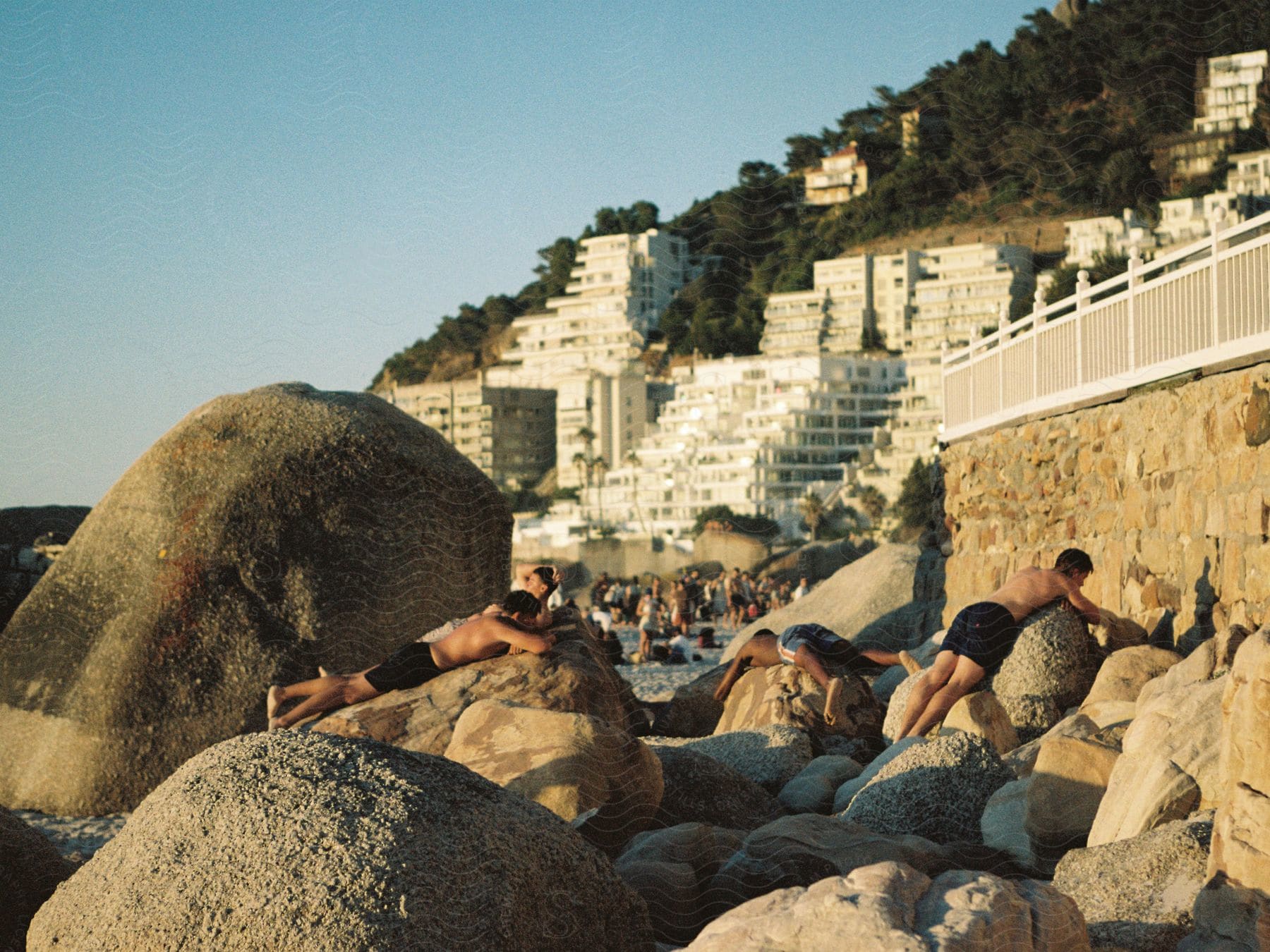 People sunbathing on large boulders with a backdrop of hillside buildings under a clear sky.