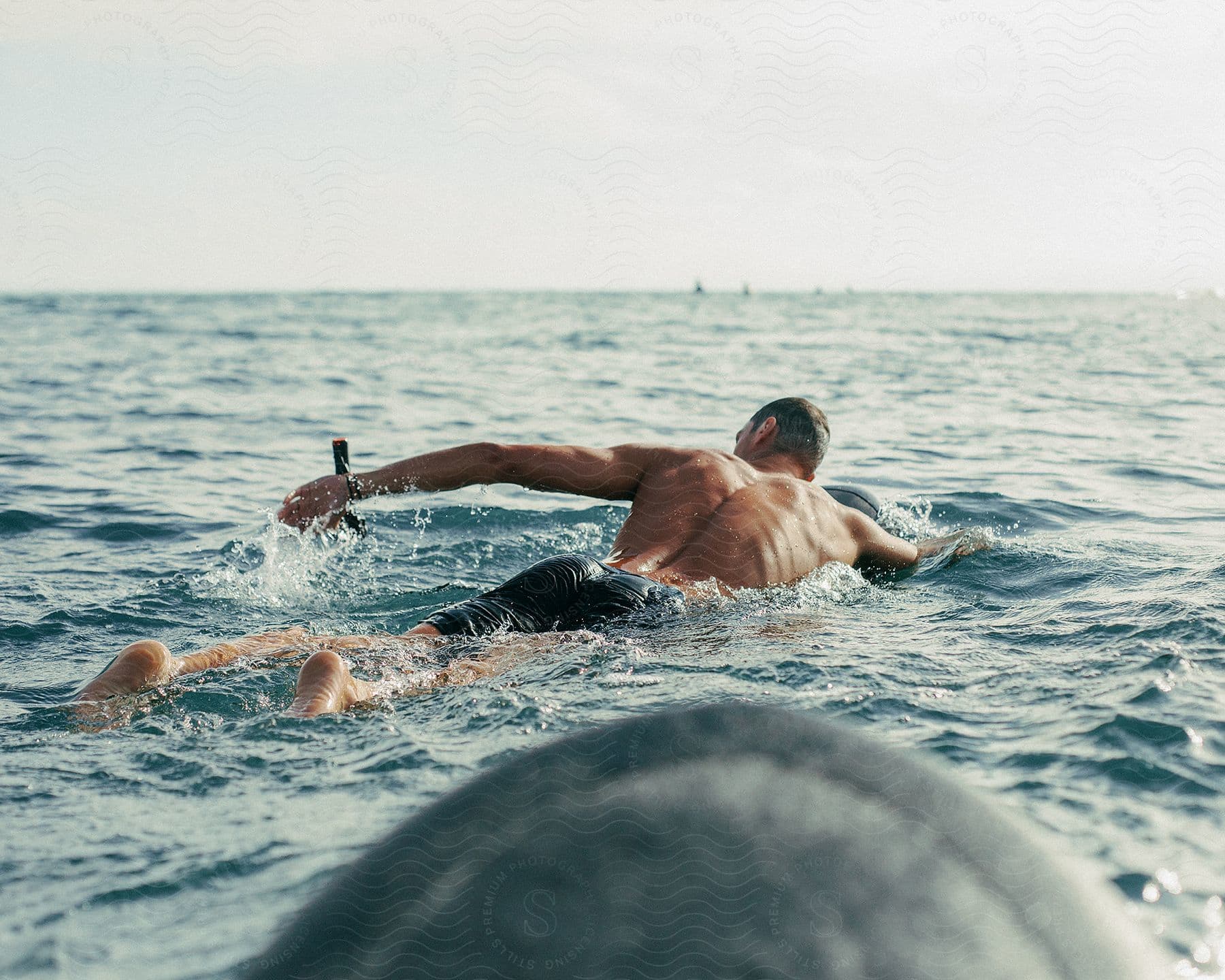 Man lying on a surfboard paddling in the ocean, view from behind with horizon visible.