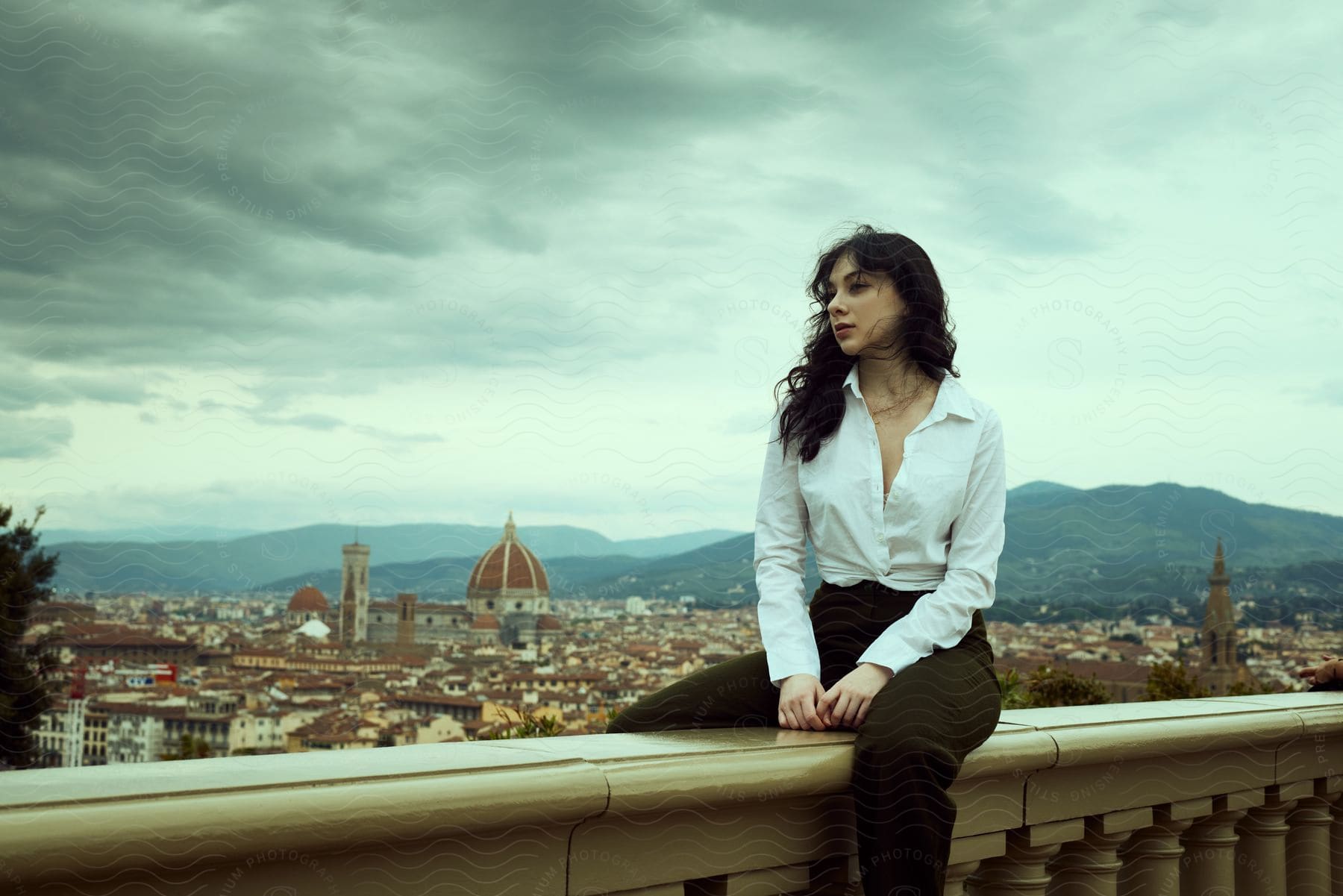 A woman is sitting on a ledge with a panoramic view of a city in the background, under a cloudy sky. The scene conveys a serene and dramatic feeling at the same time.