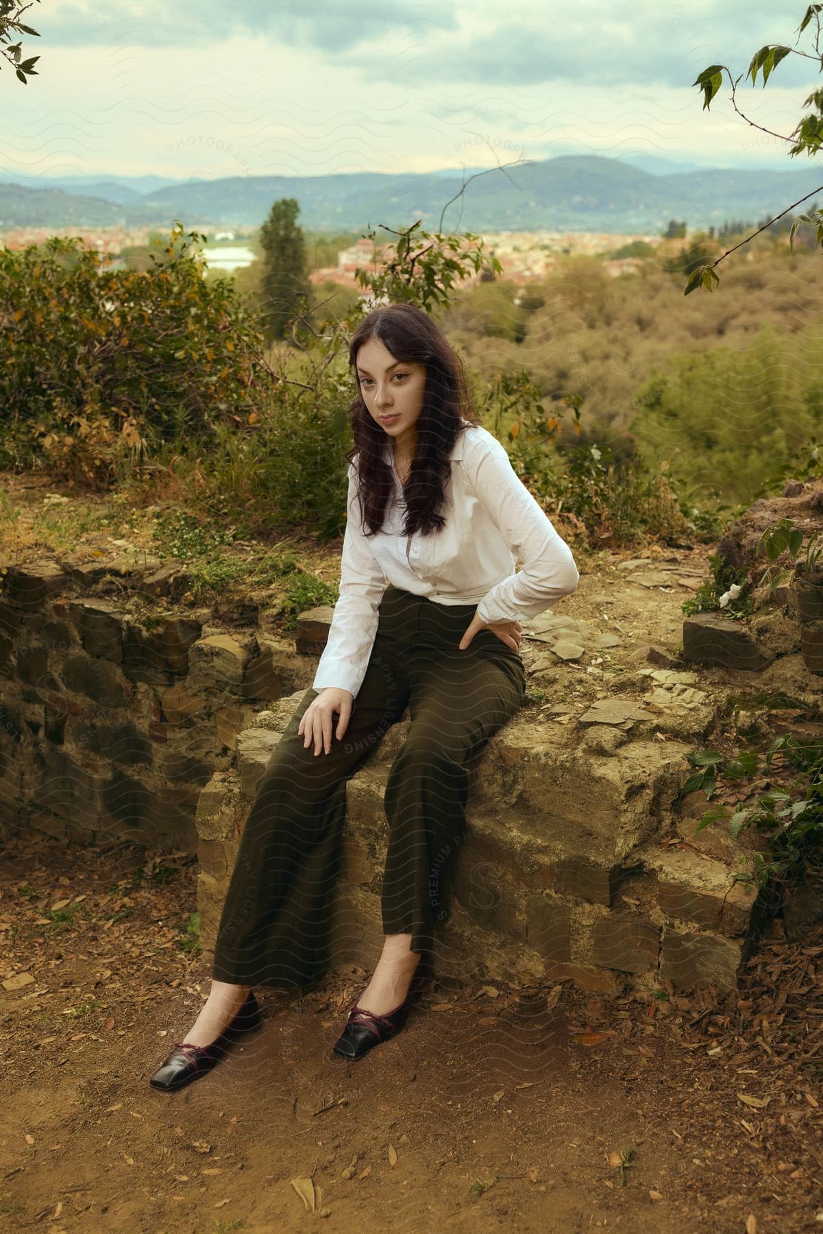 A woman wearing a white shirt, brown pants, and black shoes sits on a weathered wall in the countryside, with a town visible in the background.