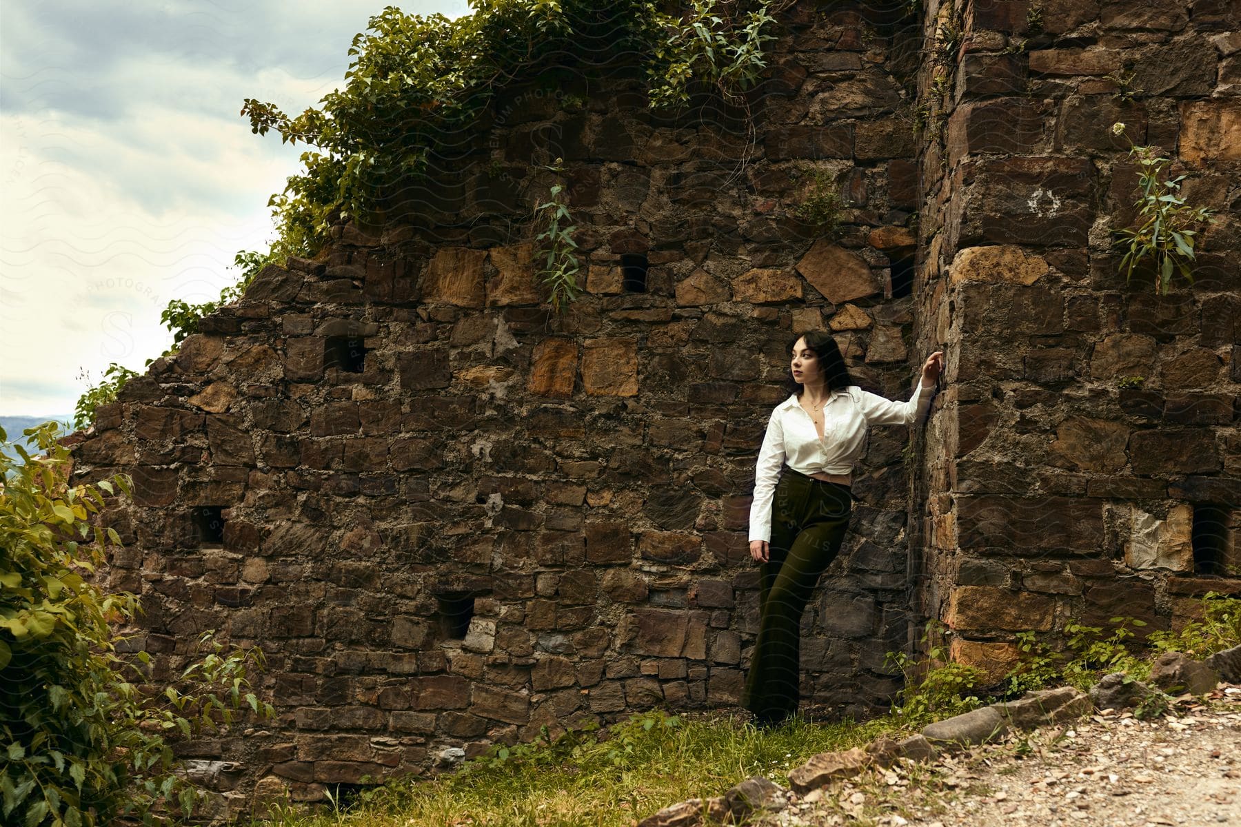 Woman model posing in pants and a white long-sleeved top next to a sloping stone wall in a natural setting.