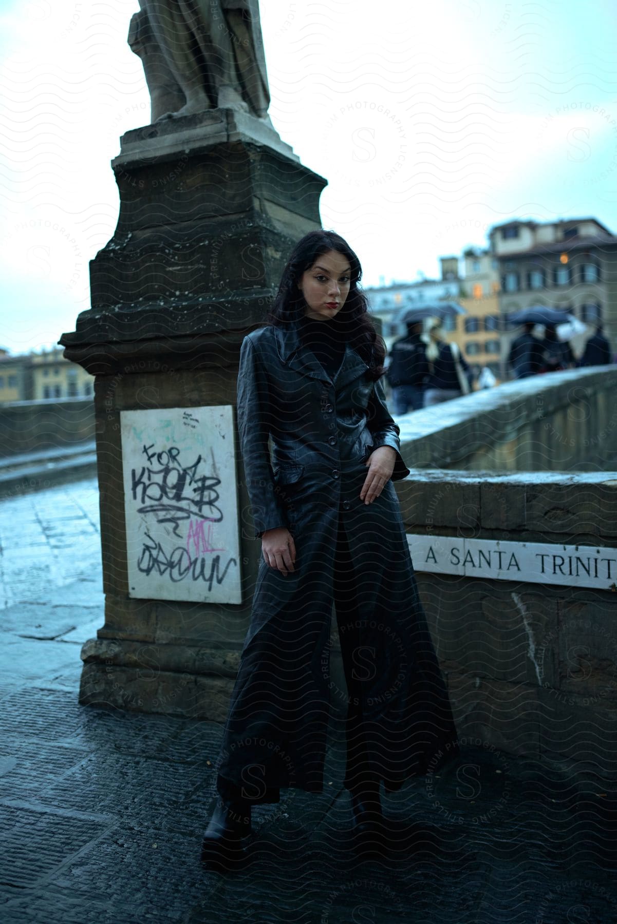 Model woman wearing a long leather overcoat modeling next to a bridge in the city