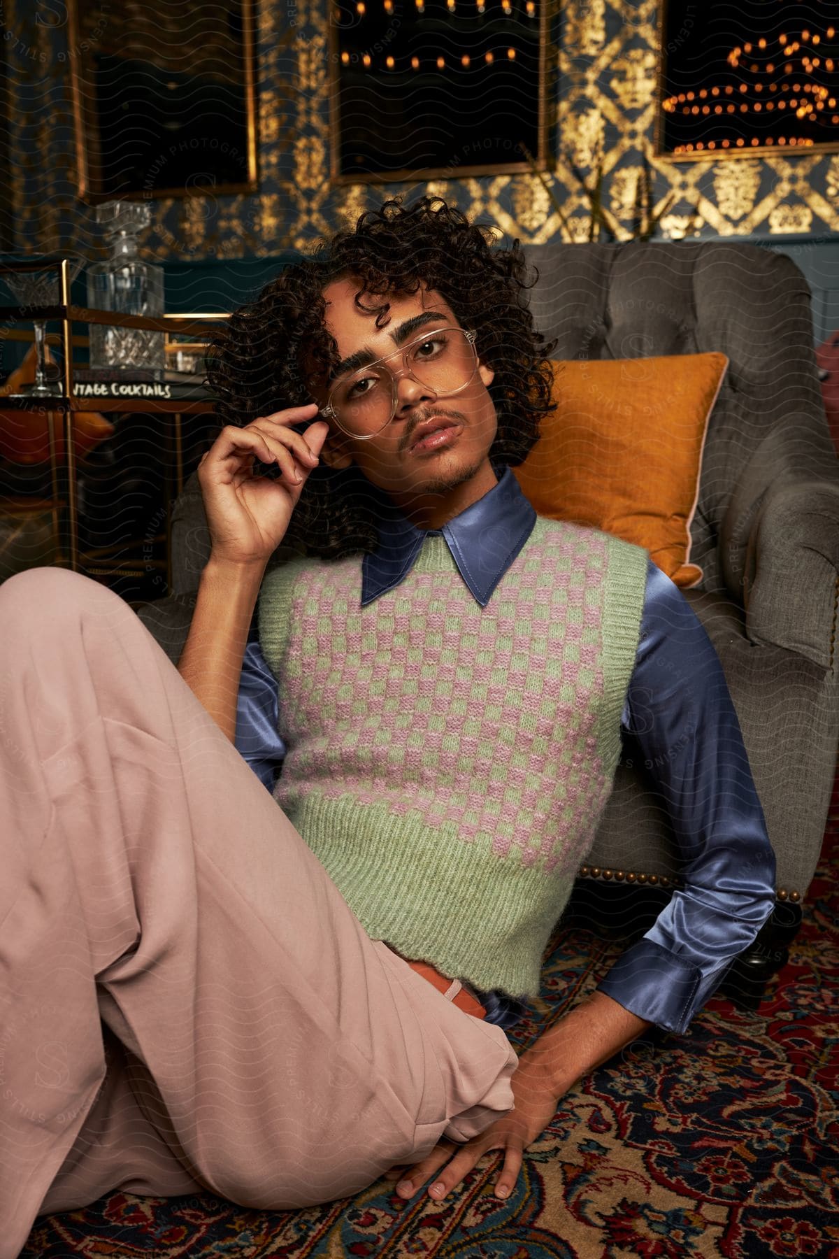 Young man with curly hair with one of his hands on his glasses posing sitting on the carpet in front of an armchair wearing pink pants and a dress shirt and sweater.
