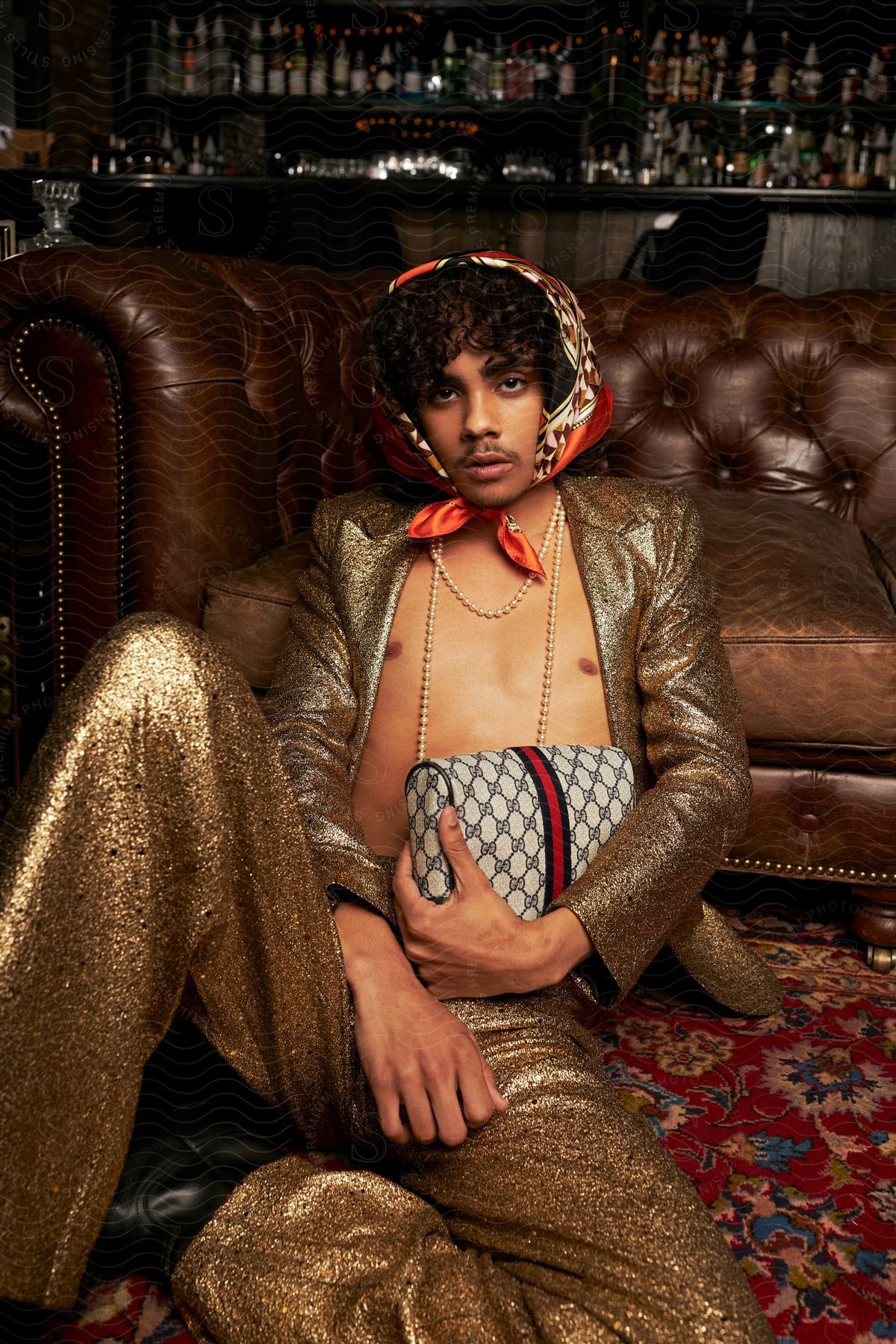 A Man With A Scarf On His Head Wearing A Sparkling Gold Open Jacket And Pants Sits On The Floor Clutching A Hand Bag