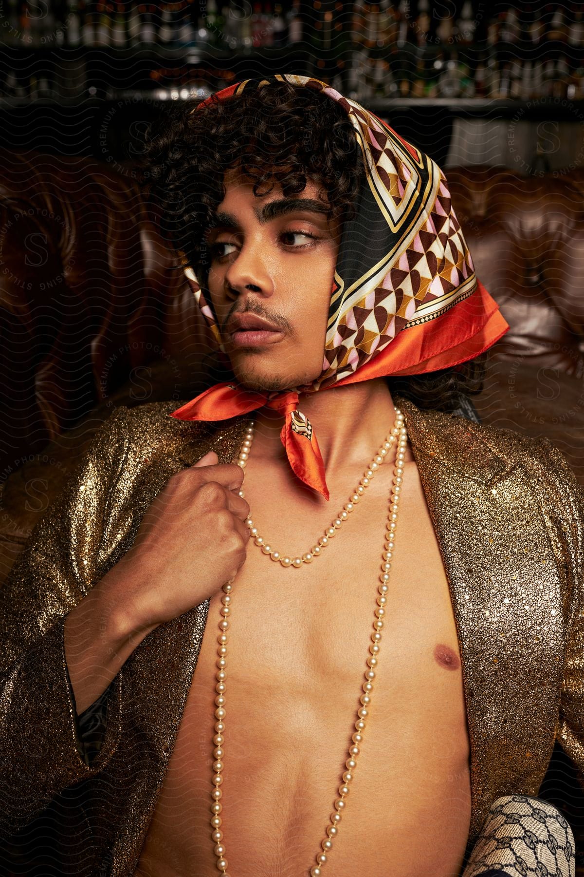 Man wearing a head scarf and an open gold jacket with a bare chest as he holds the pearls around his neck and looks to his side