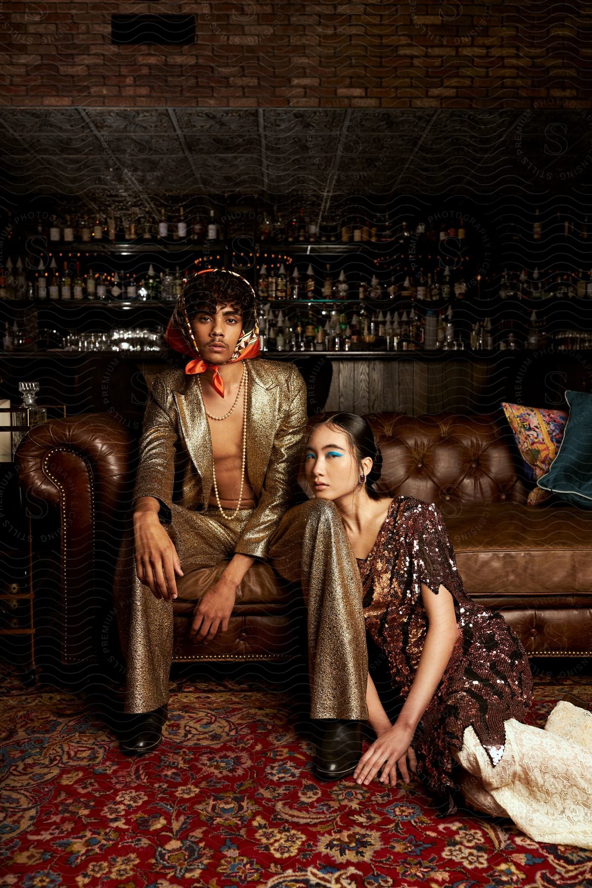 A man and a woman are sitting on a brown leather sofa, wearing light-colored clothes, in front of a bar.