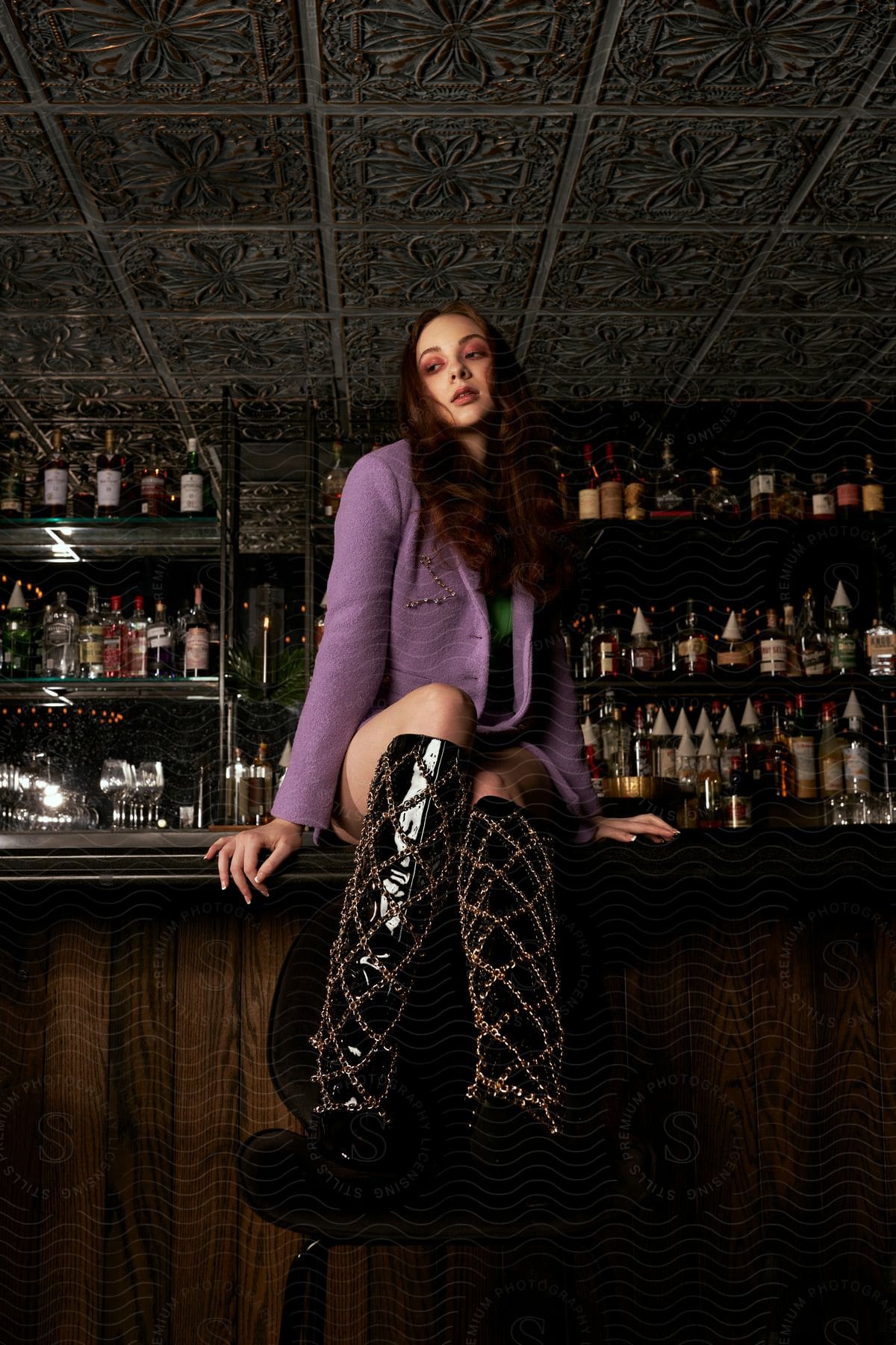 Woman wearing elaborately detailed tall boots sits on counter in bar.