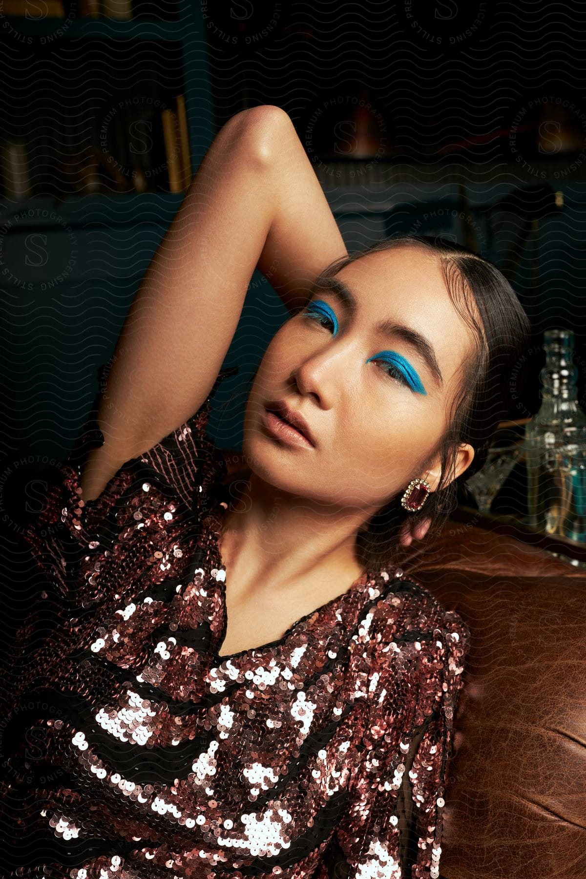 A portrait of an Asian woman with blue eye makeup and wearing a dress with small bright red details and she has her hand on the back of her head