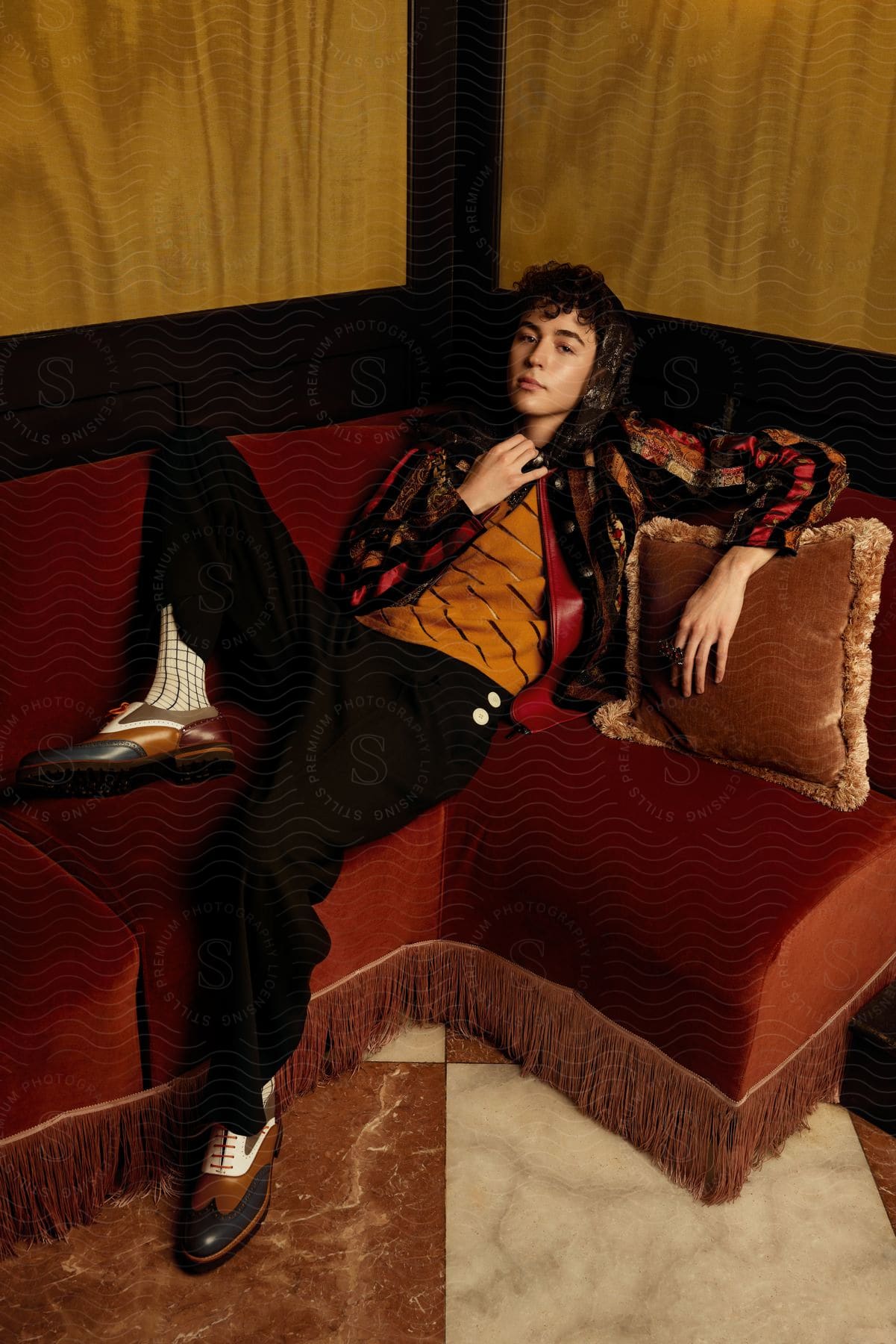 A person lounges elegantly on a velvet sofa, wearing a patterned jacket, yellow vest, and checkered shoes, evoking a vintage, luxurious style.