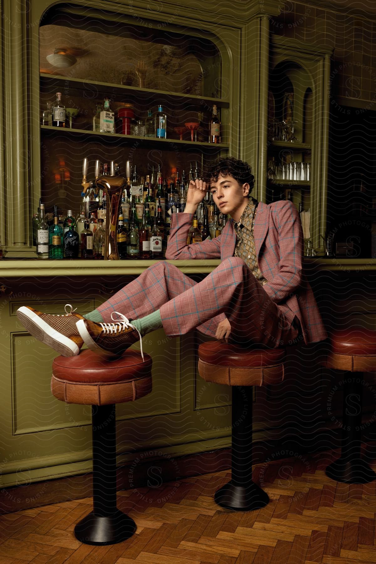 A young man sitting on a bar stool with his feet on another, at the counter of an old style bar.