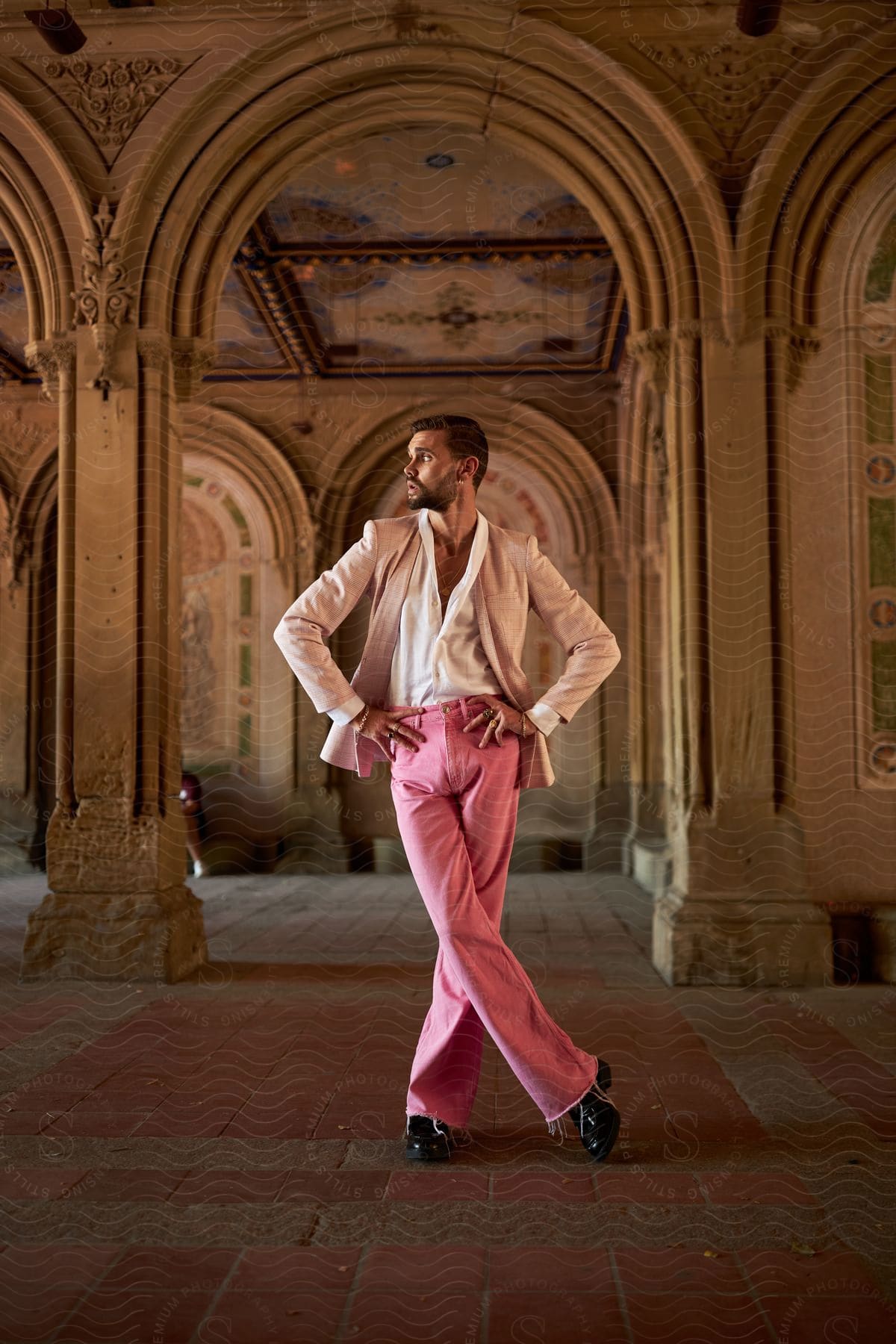 A man wearing pink pants and a lighter blazer has his hands on his waist while looking to his right side in an environment with rustic architecture