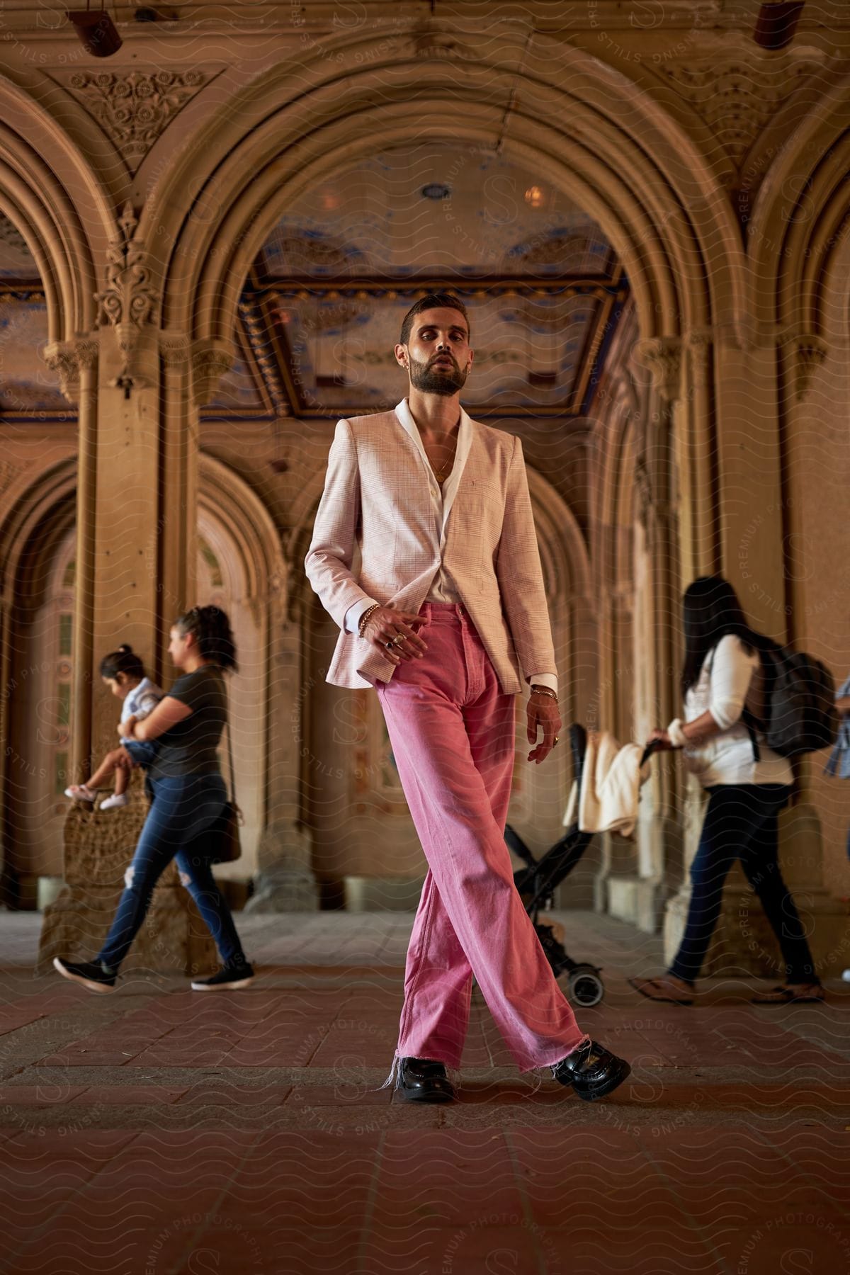 Man posing in beige jacket and pink trousers in an ornate corridor with people in the background.