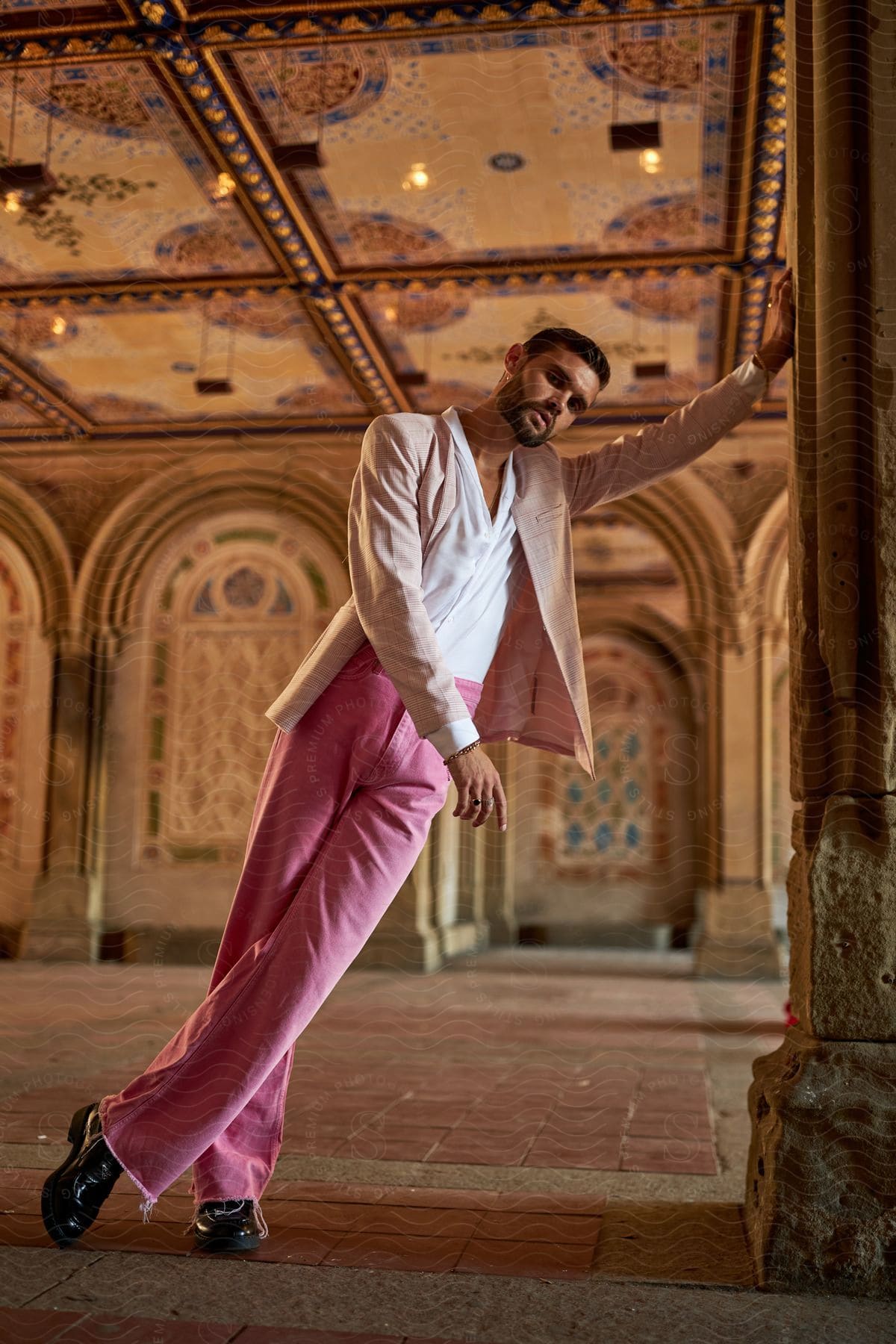 Man wearing a blazer and pink pants leans against a pillar in an ancient building