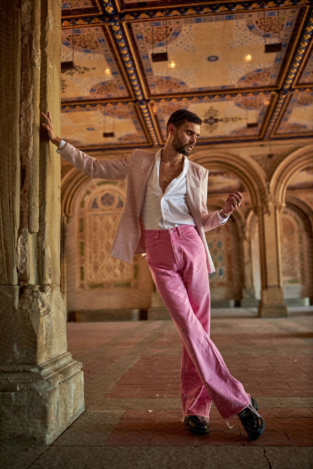 Man in pink trousers and beige blazer leaning against stone pillar, ornate ceiling overhead.