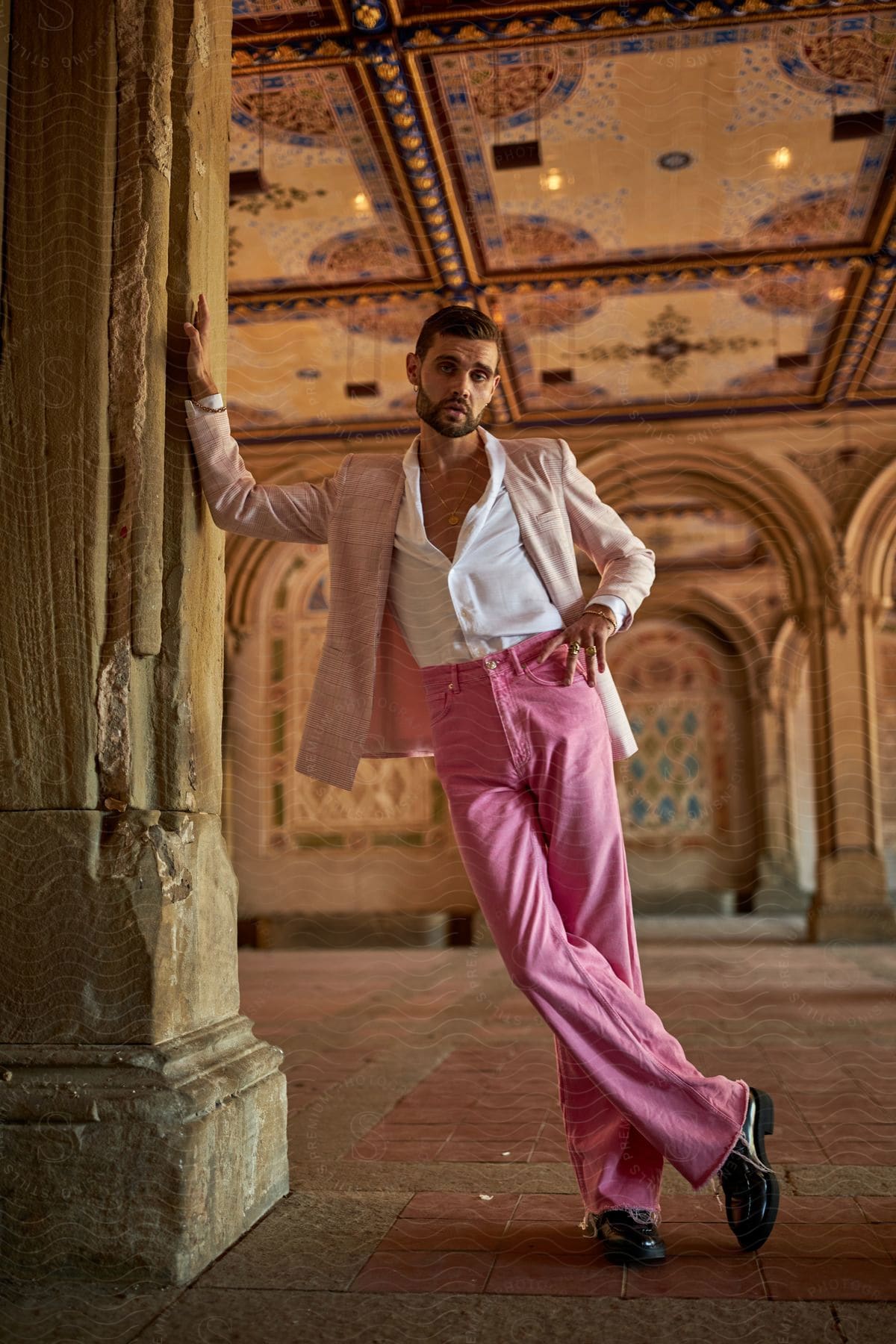 A man in pink pants and wearing a blazer is striking a pose while placing his hand on an ancient architectural pillar