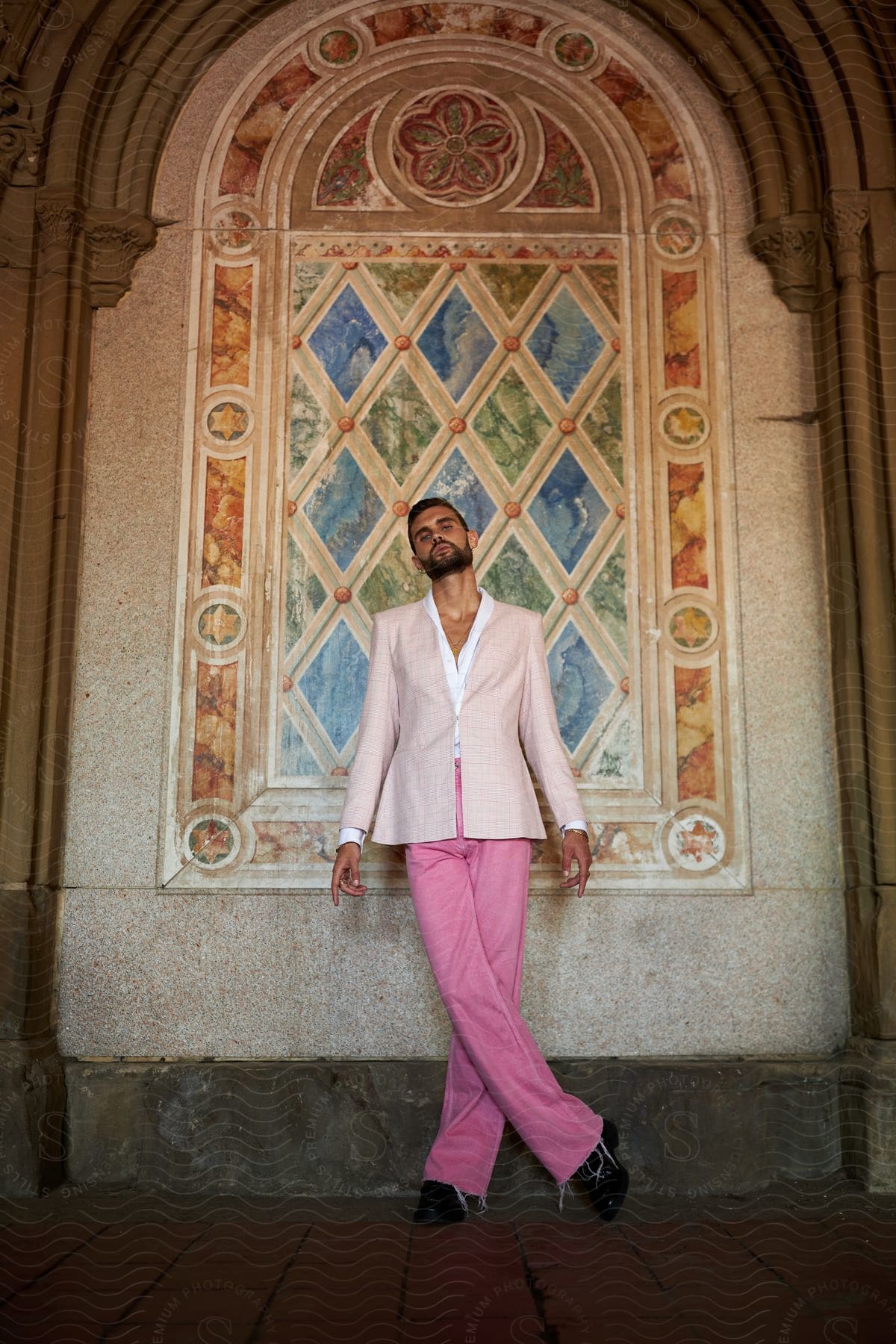 A male model in pink pants and tan jacket posing in front of a painted false window.