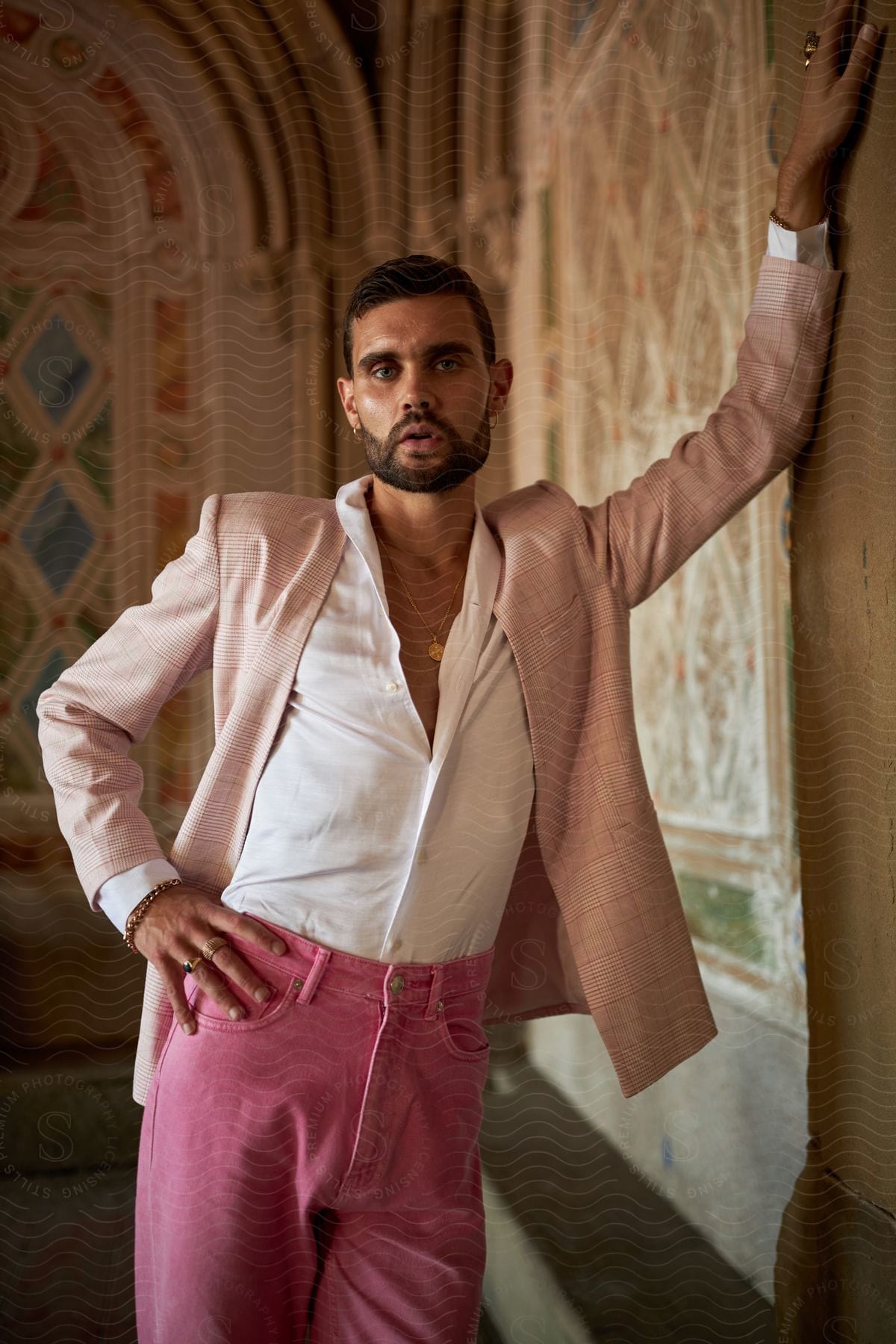 A man poses elegantly against an ornate wall, wearing a light pink blazer, white shirt and pink pants.