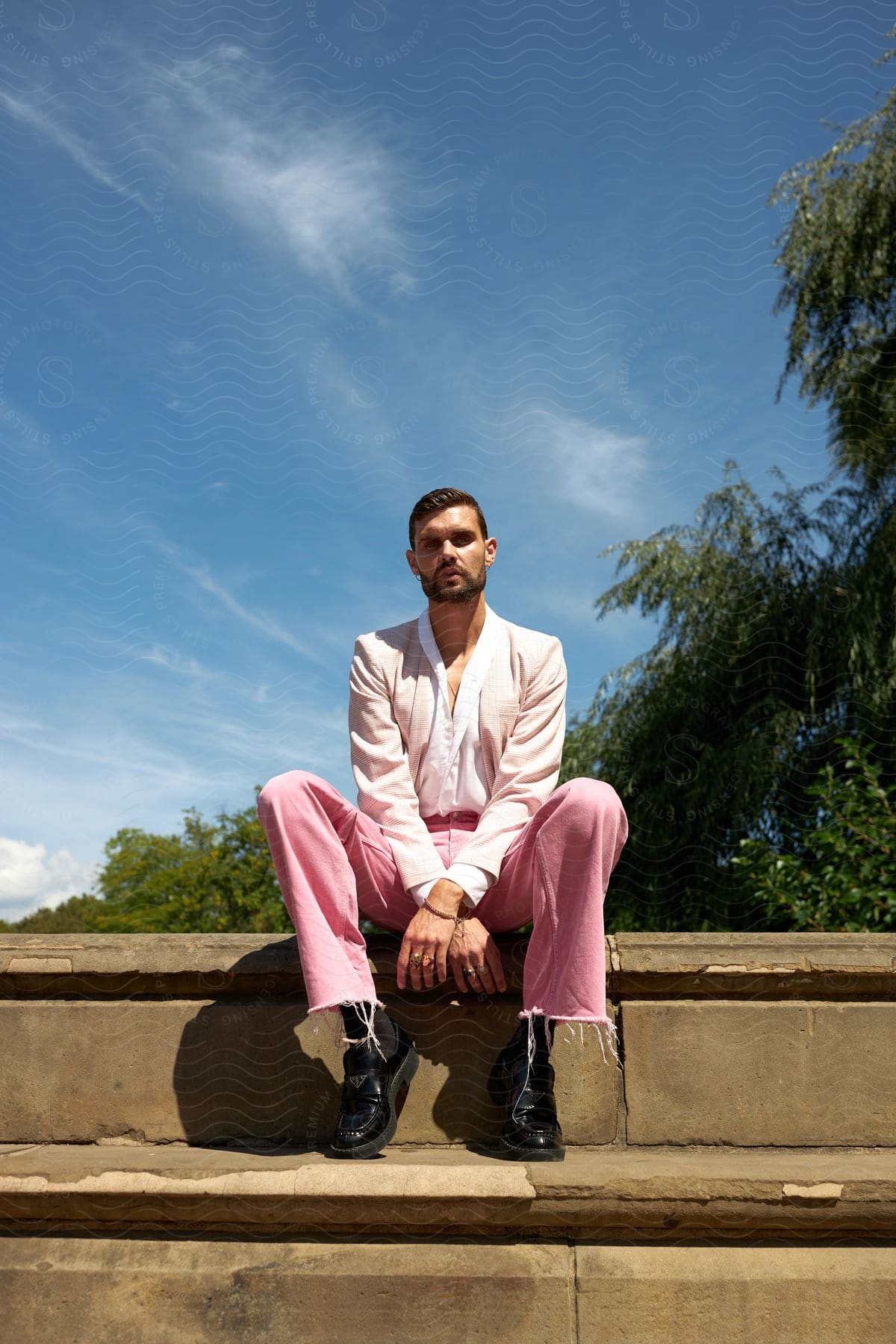 A barded man in white shirt and pink pants sits on stone step in front of trees.