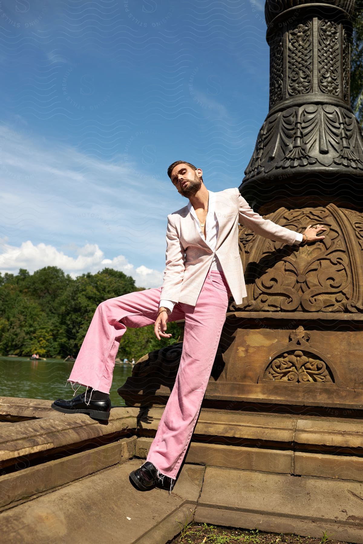 Model man posing in a blazer and a pink sidewalk next to a monument on the edge of a lake.