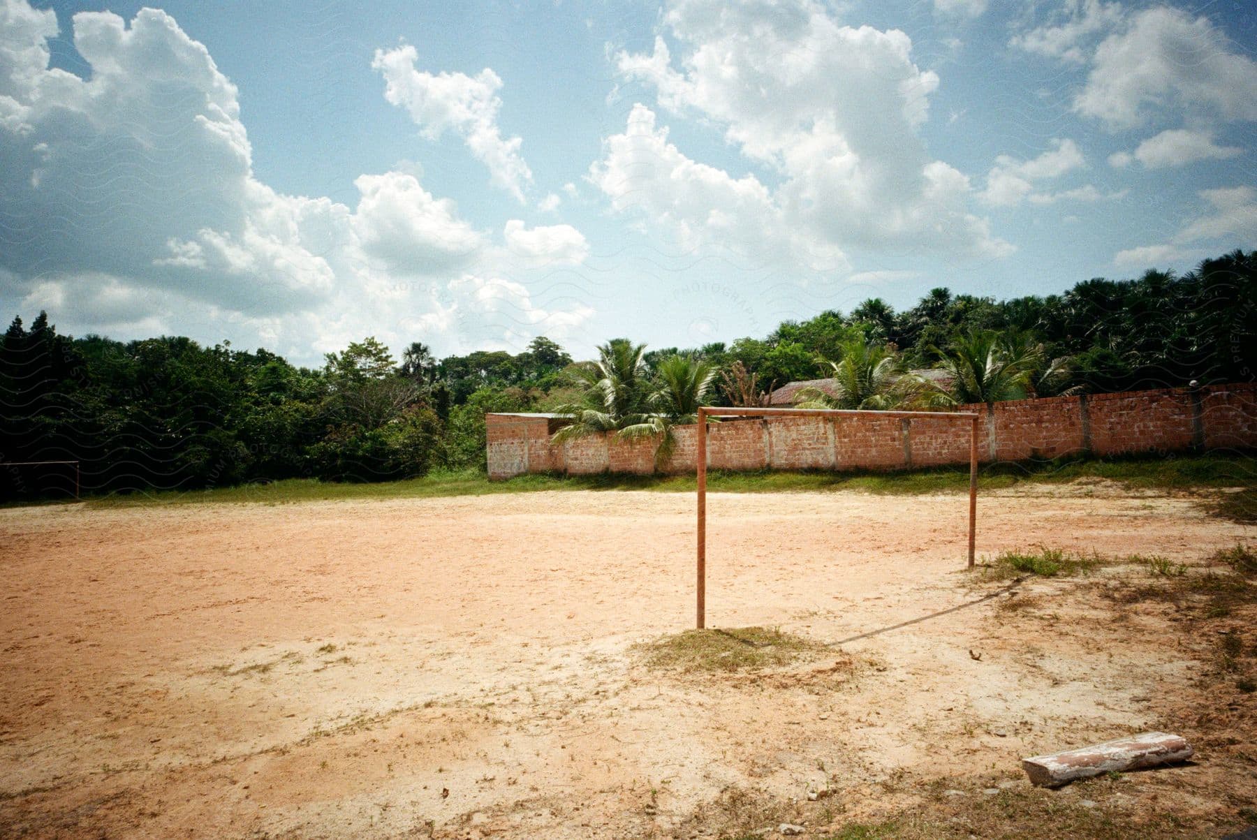 An outdoor dirt soccer field, with the net missing. The soil of the field is uneven and worn, with undergrowth growing in some spots.