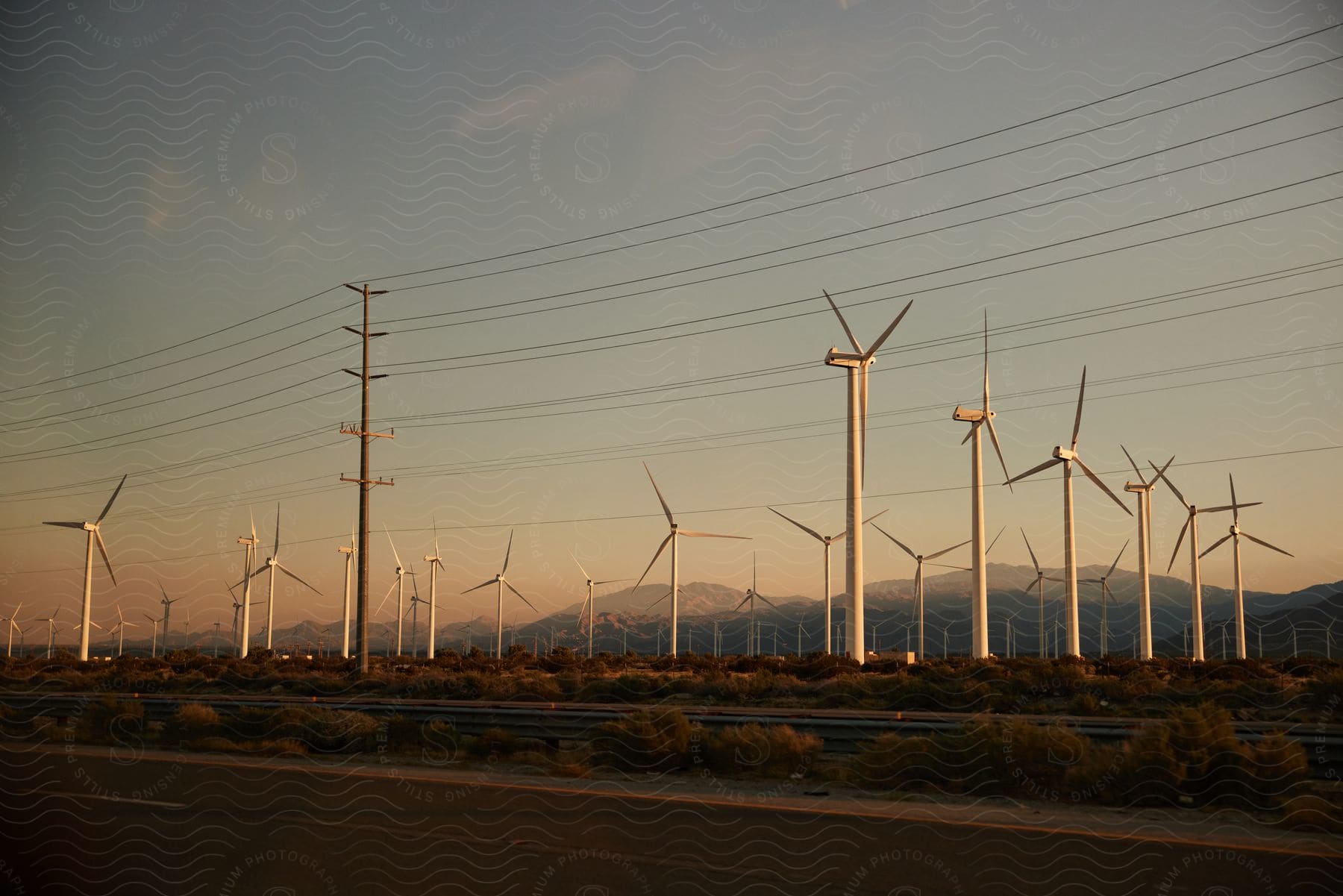 Wind turbines stand on a wind farm near utility wires with mountains in the distance