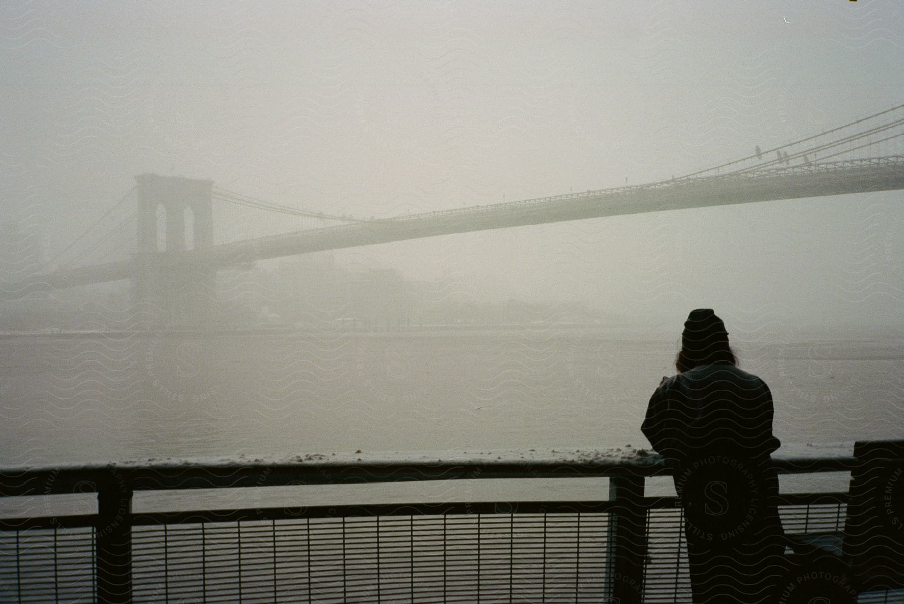 Person wearing a hat and black overcoat leans against the railings, observing the Brooklyn Bridge enveloped in fog.