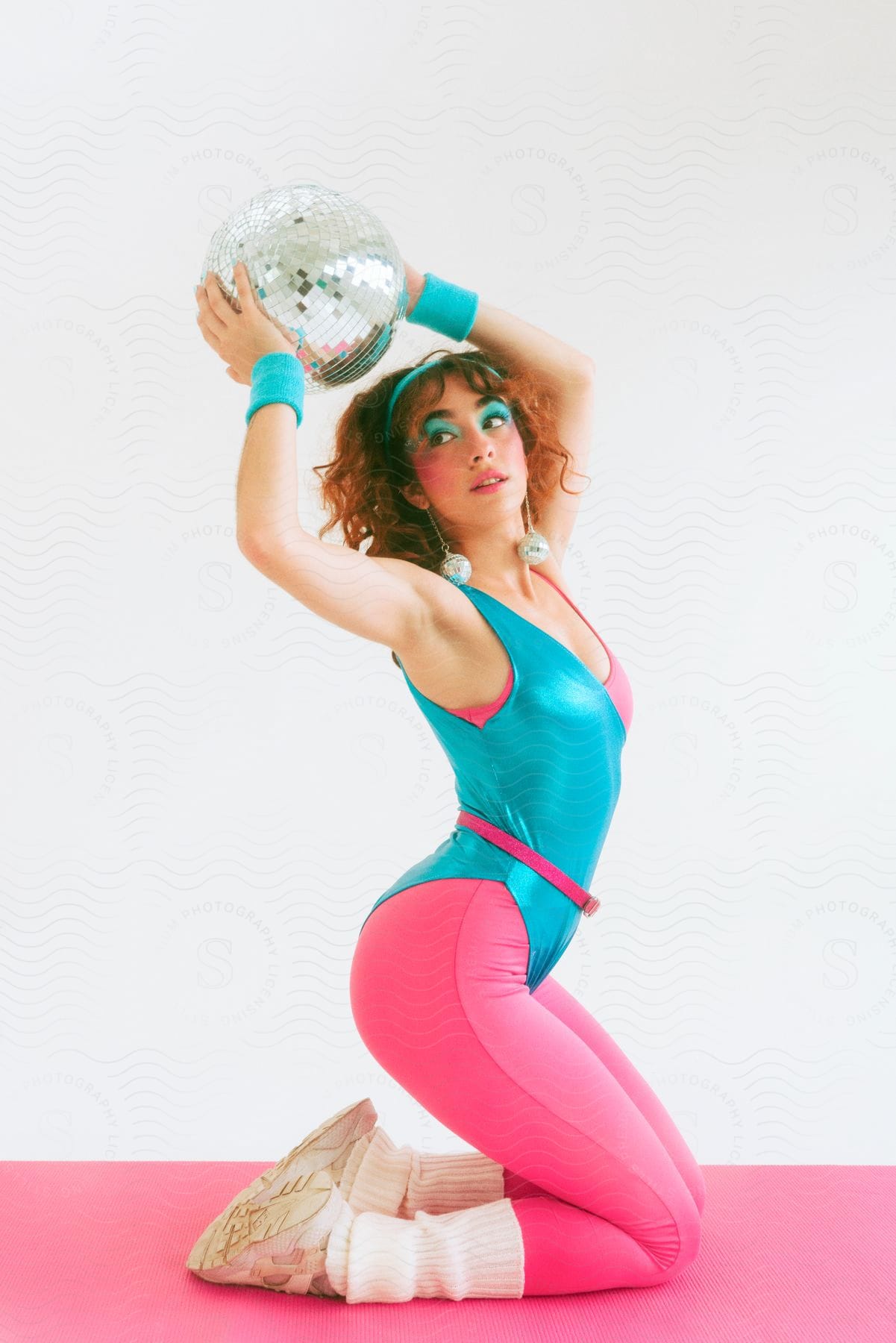 Woman in a teal leotard with pink leggings and leg warmers holding a disco ball over her head, posed on a pink mat against a white background.