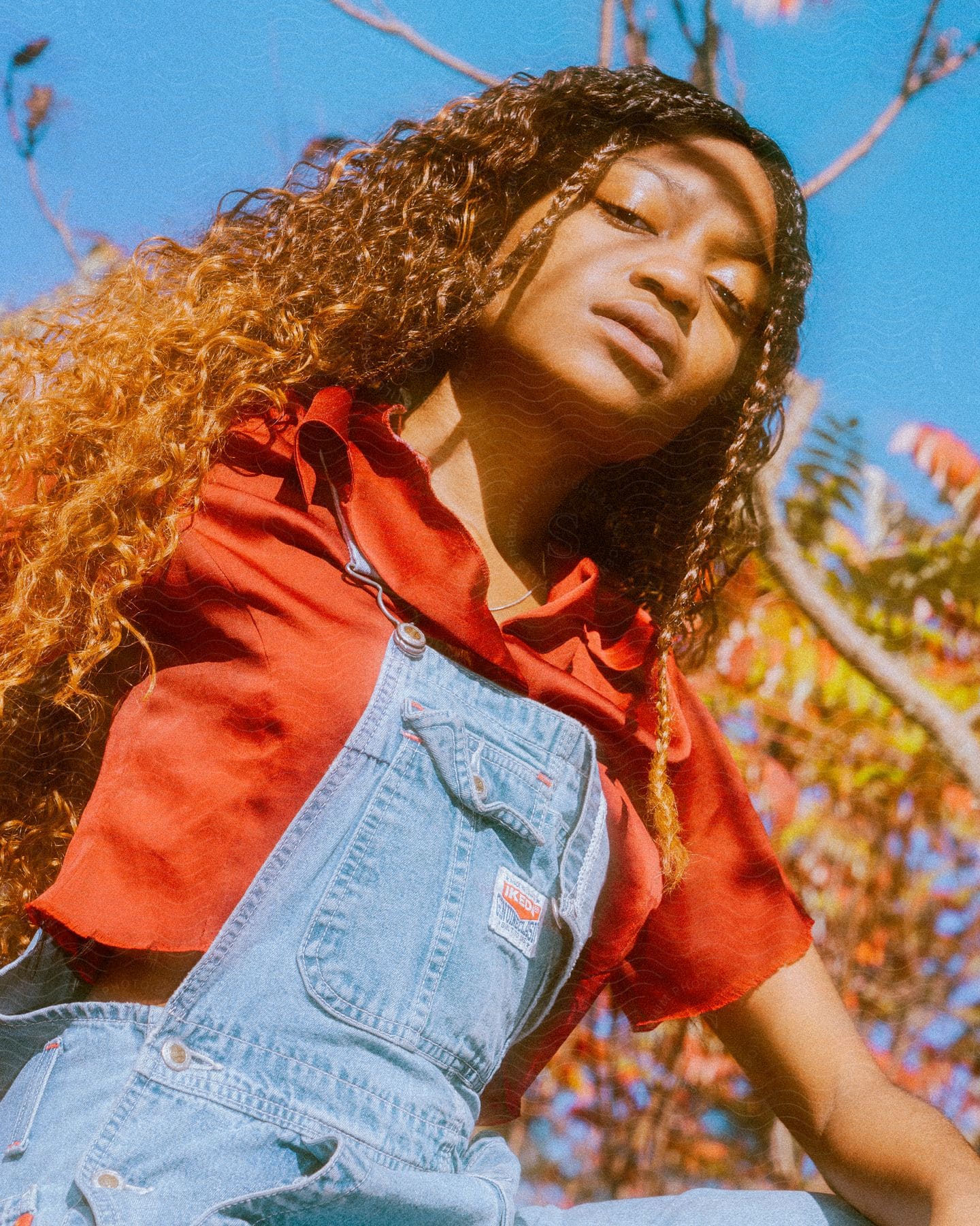 A young black teenage girl with curly brown braided hair, dressed in a red shirt, pose outdoors under a tree.