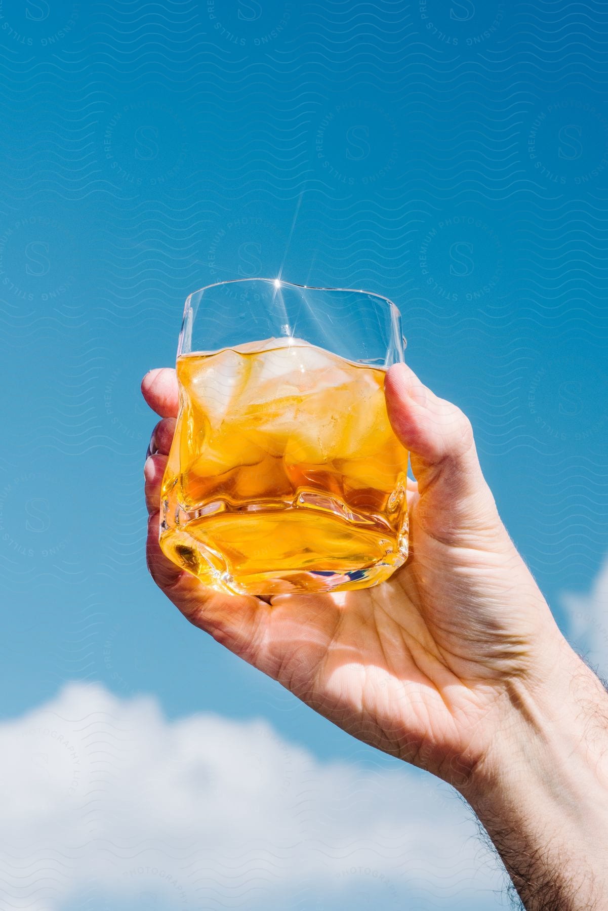 Right hand holds Amber spirit beverage in short glass with ice against blue sky background
