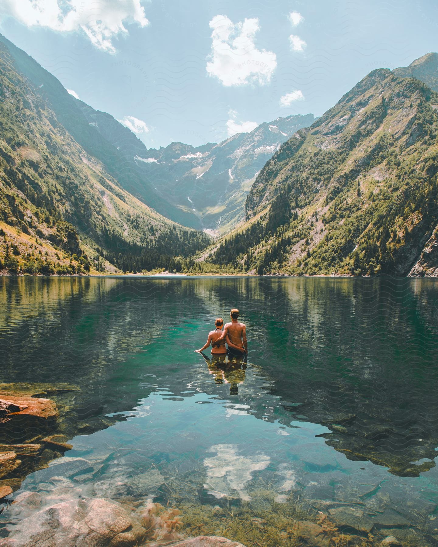 Two people together standing in a clear mountain lake with green slopes and peaks in the background.