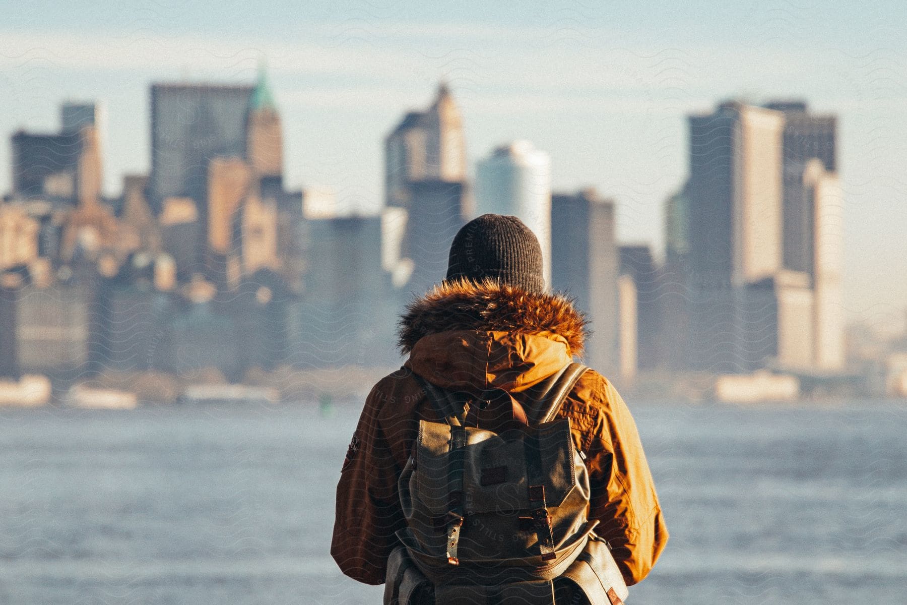 A person wearing a hooded winter coat and backpack looking out over the water at city skyscrapers